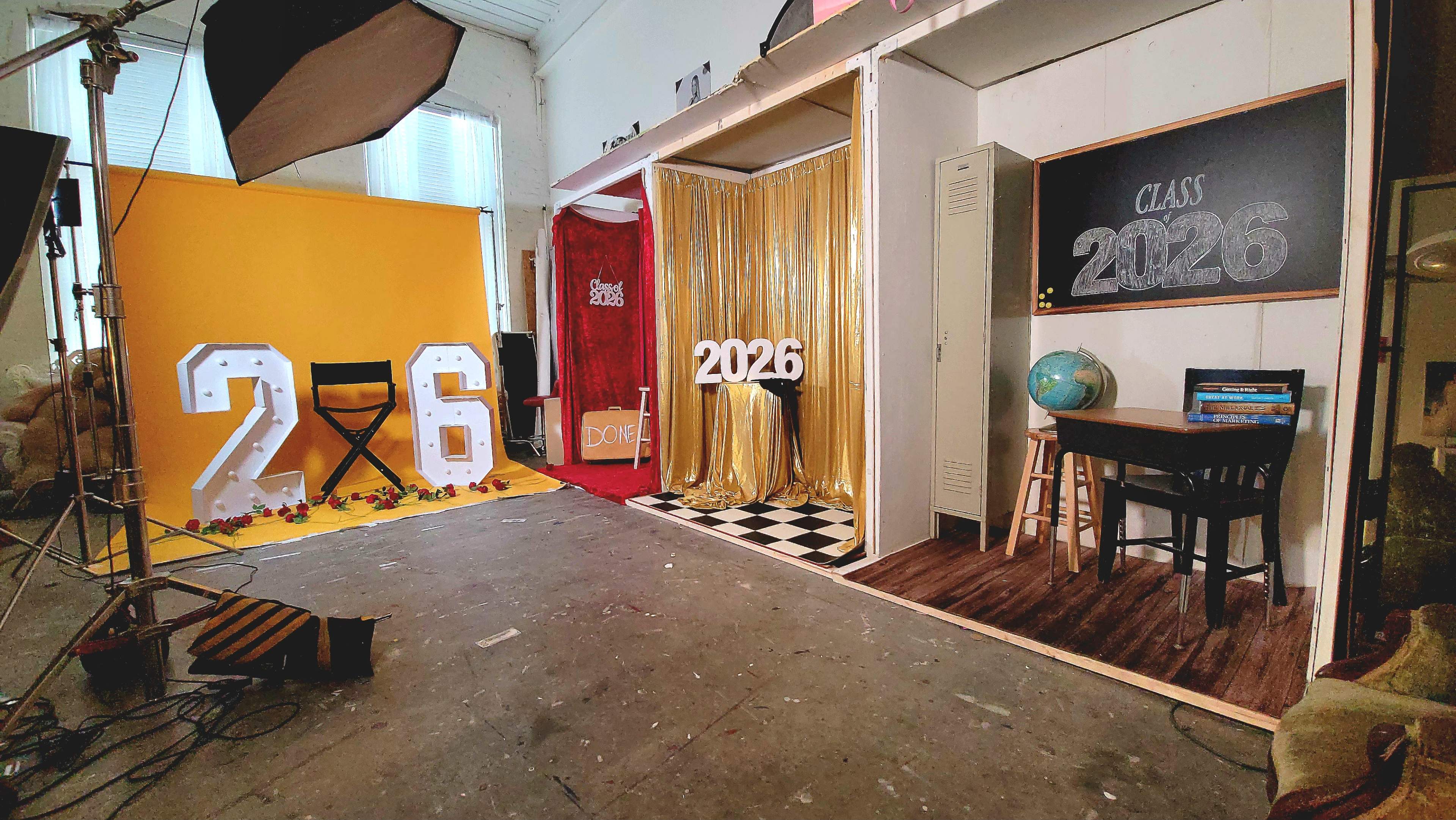The image shows a photo studio setup for a graduation-themed shoot, featuring the numbers "2026," gold curtains, a chalkboard, a globe, and a wooden chair.