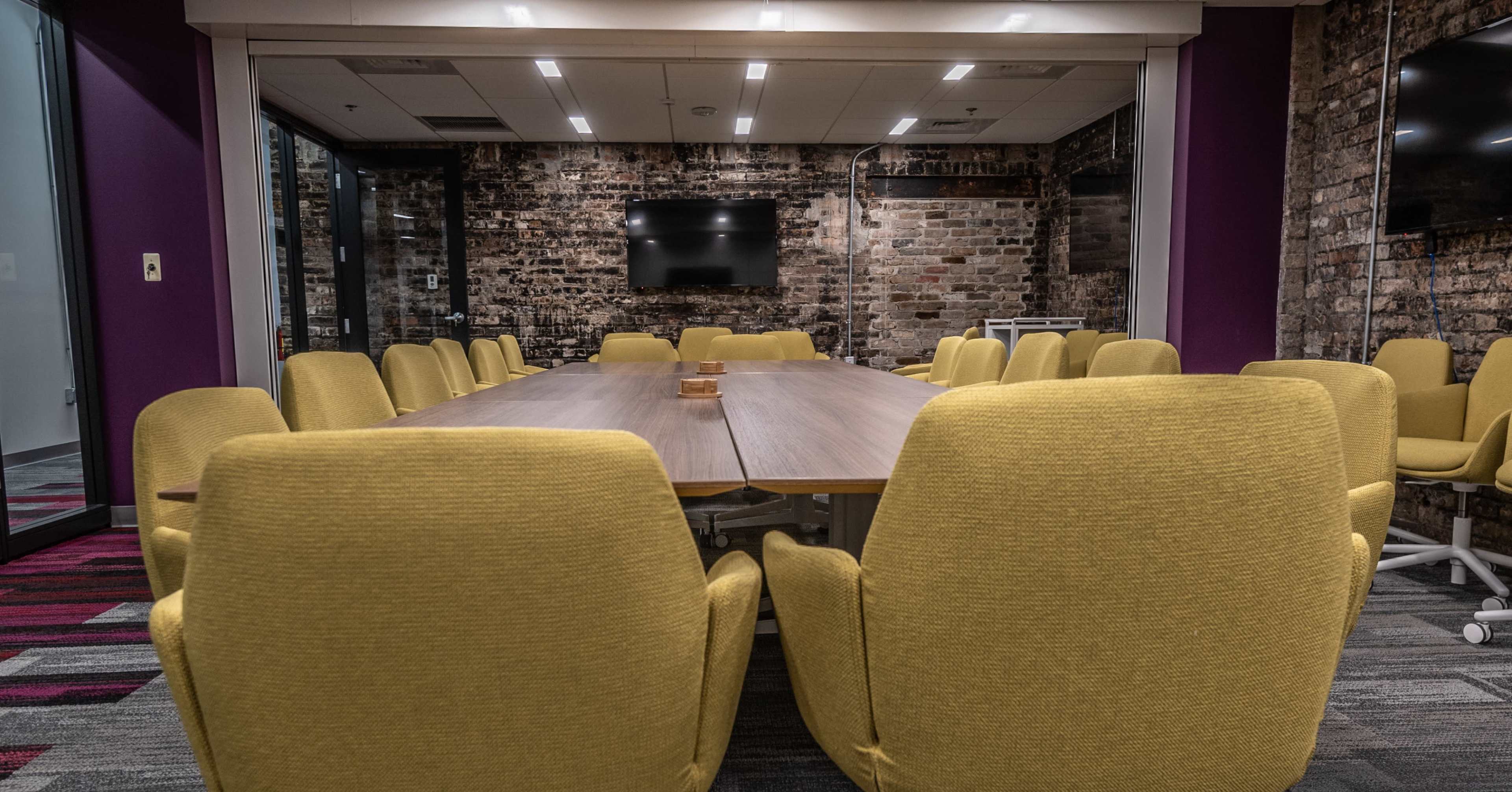 A conference room features a long table surrounded by yellow chairs, with exposed brick walls and modern technology visible.