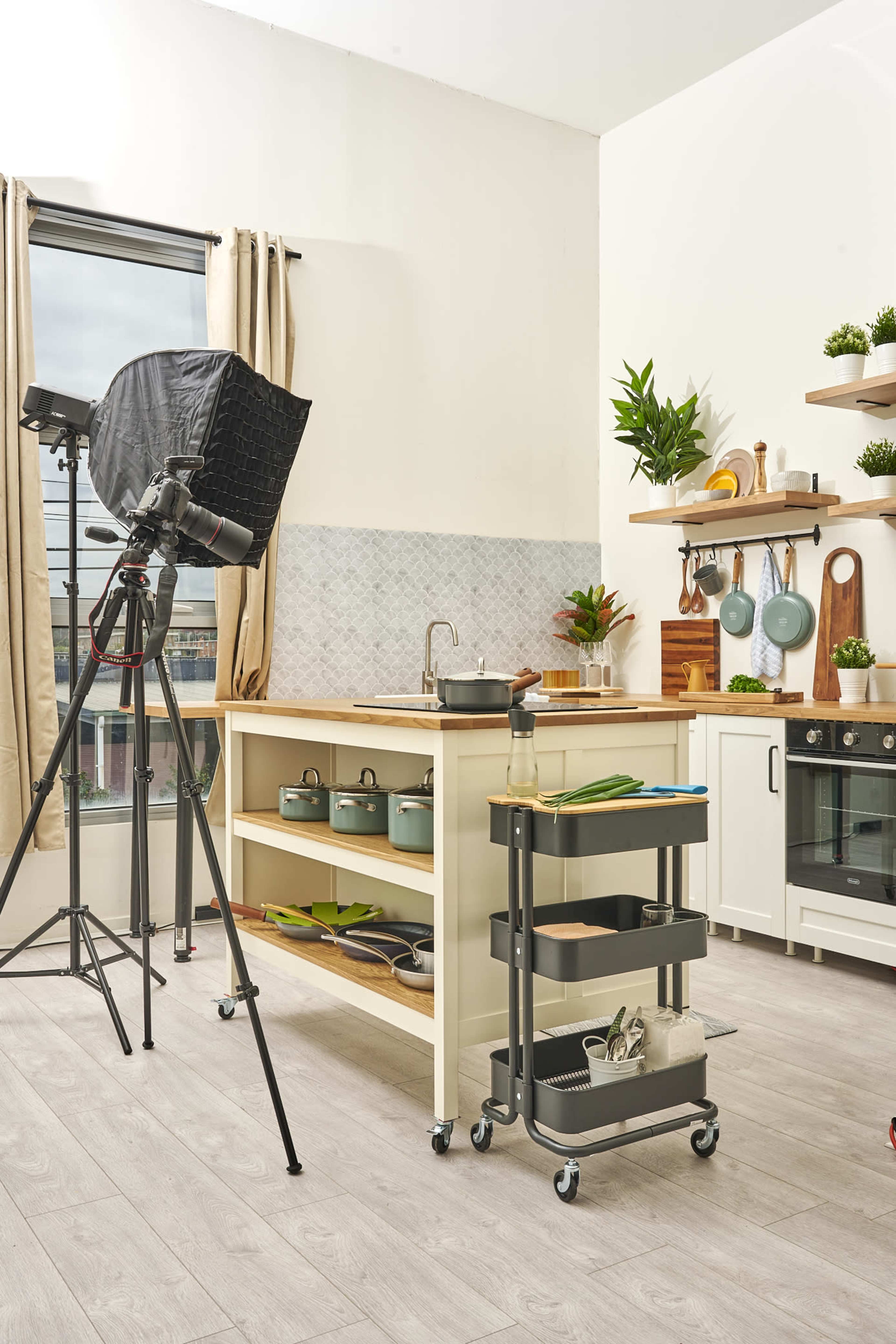 A well-lit kitchen scene features a wooden island with cooking utensils and plants, accompanied by a camera setup on a tripod for filming.