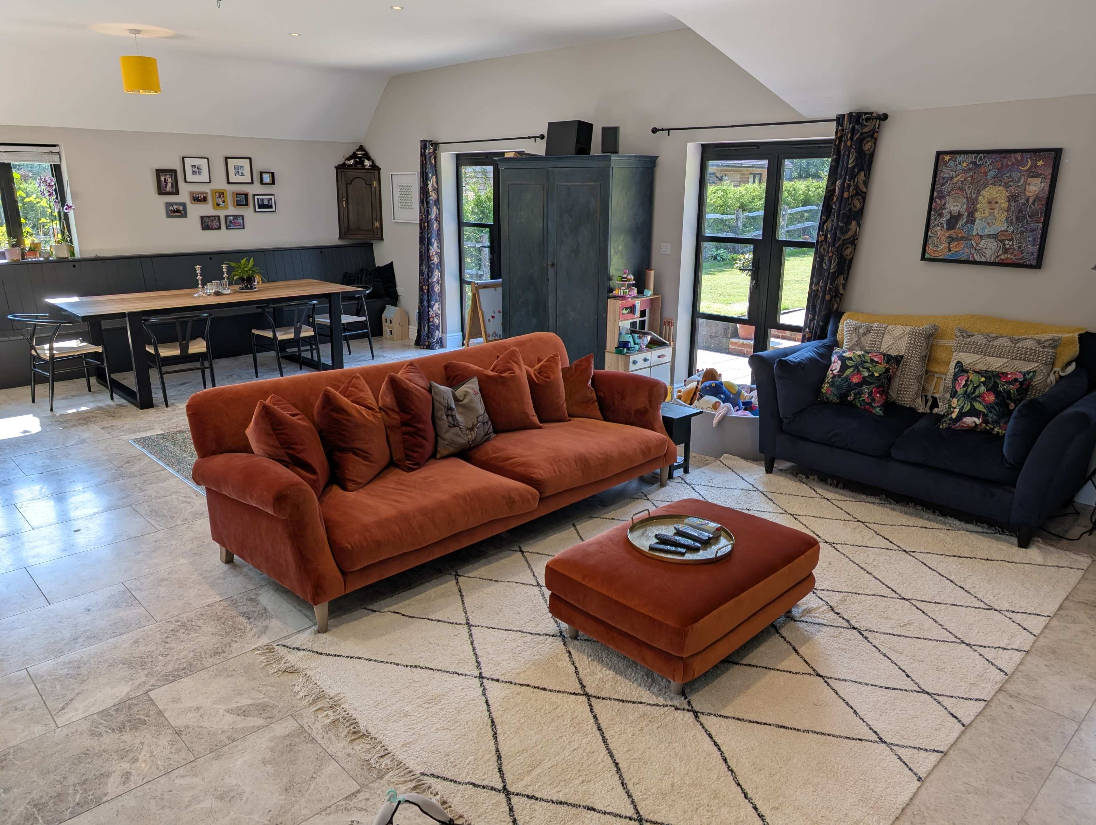 The image shows a spacious living area featuring an orange sofa, a dark blue couch, a rectangular dining table, and a large window overlooking a garden.