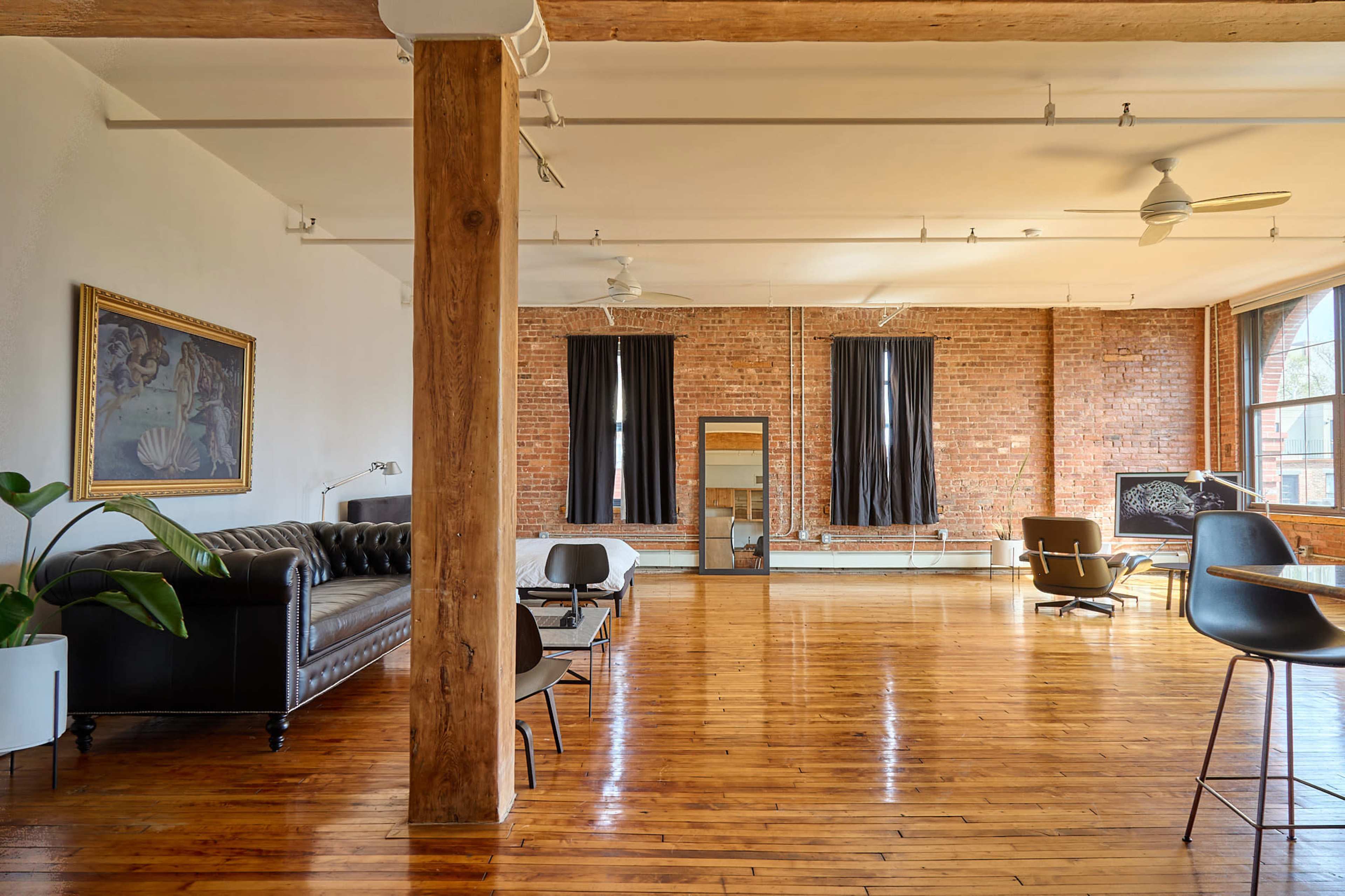 The image shows a spacious loft with hardwood floors, exposed brick walls, and various seating arrangements, including a leather sofa and modern chairs.