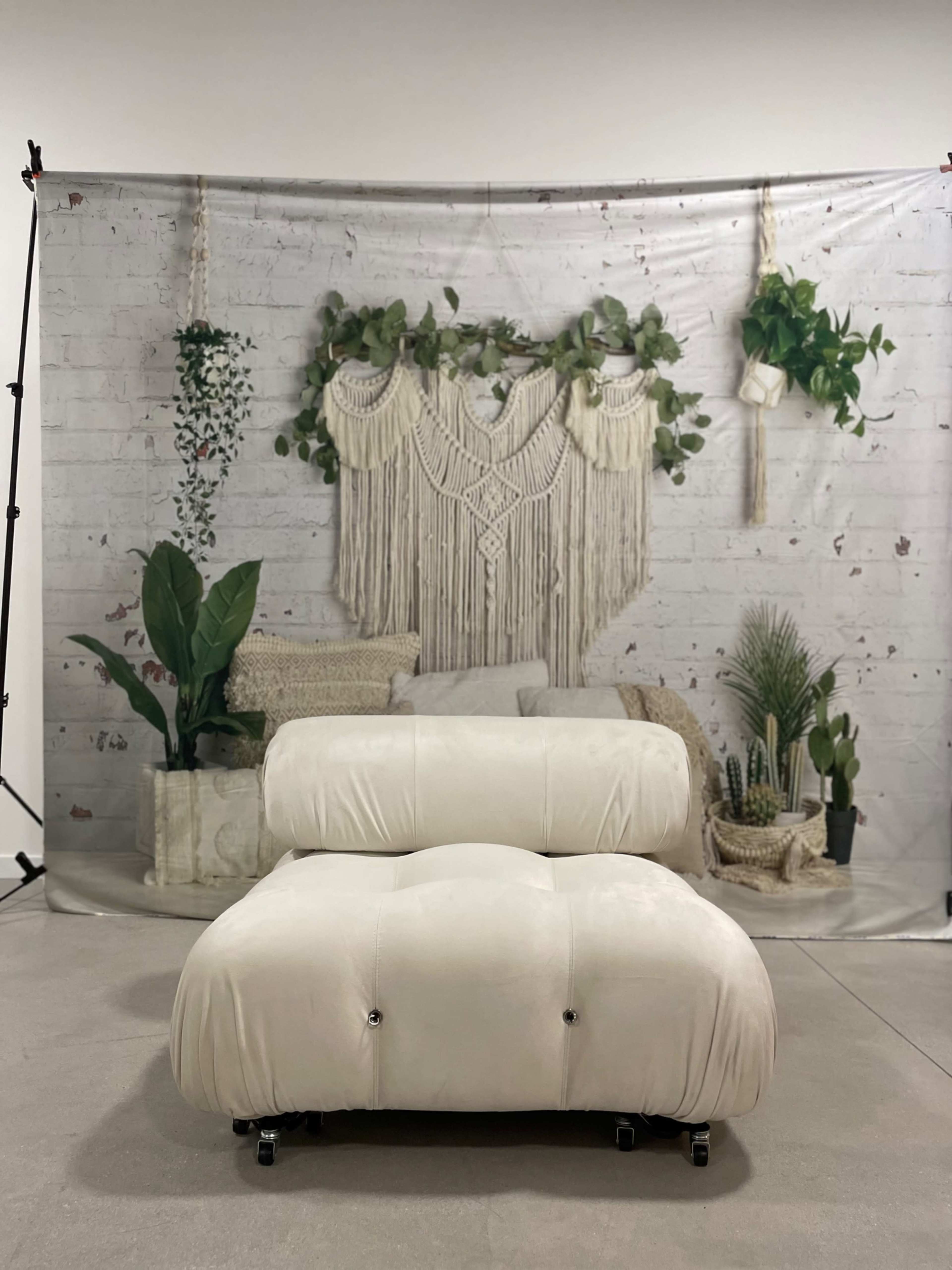 A cream-colored chair in front of a backdrop featuring a macramé wall hanging and various potted plants.