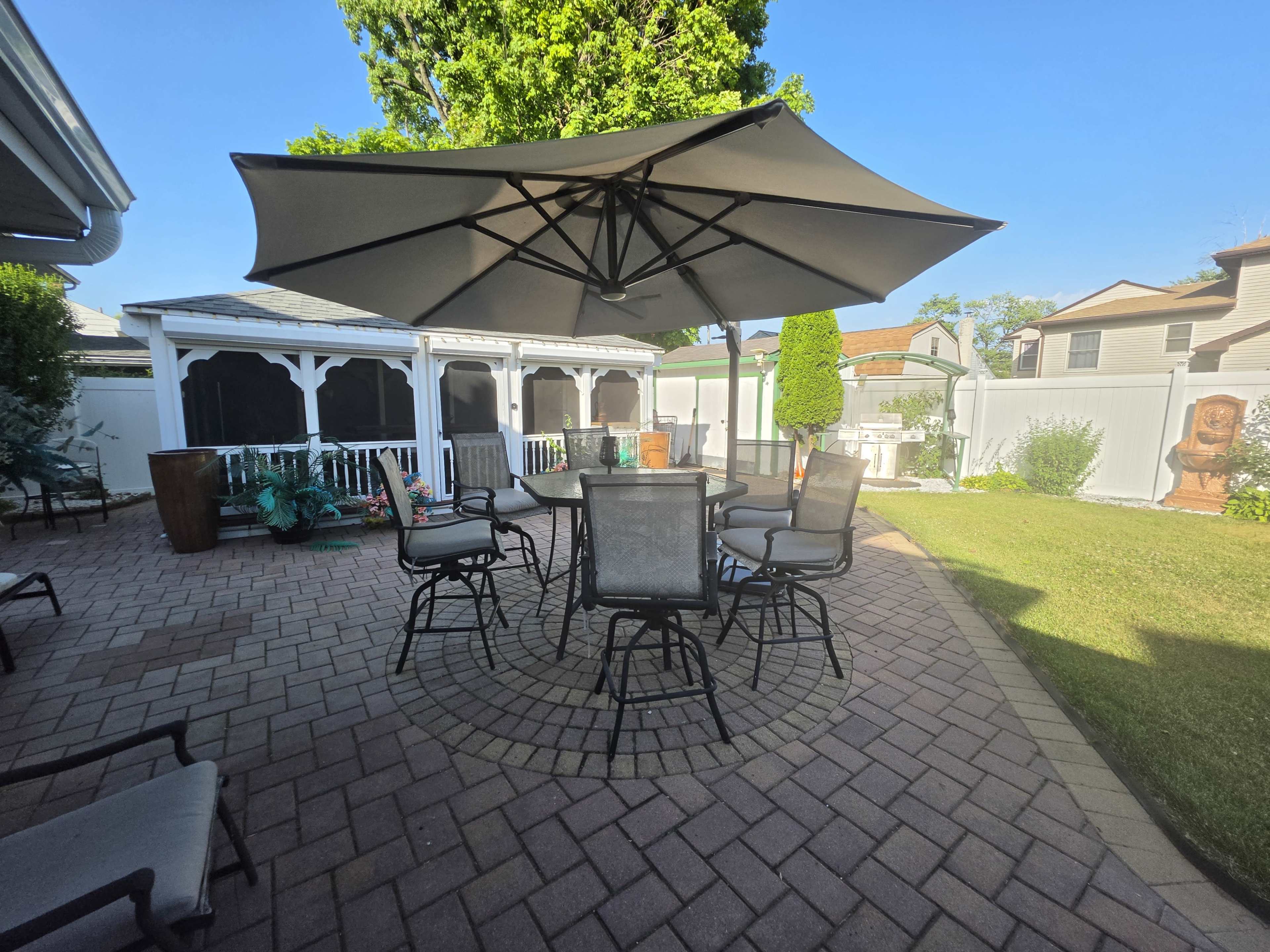 A patio area features a large umbrella over a circular table surrounded by several chairs, with a gazebo and landscaped greenery in the background.