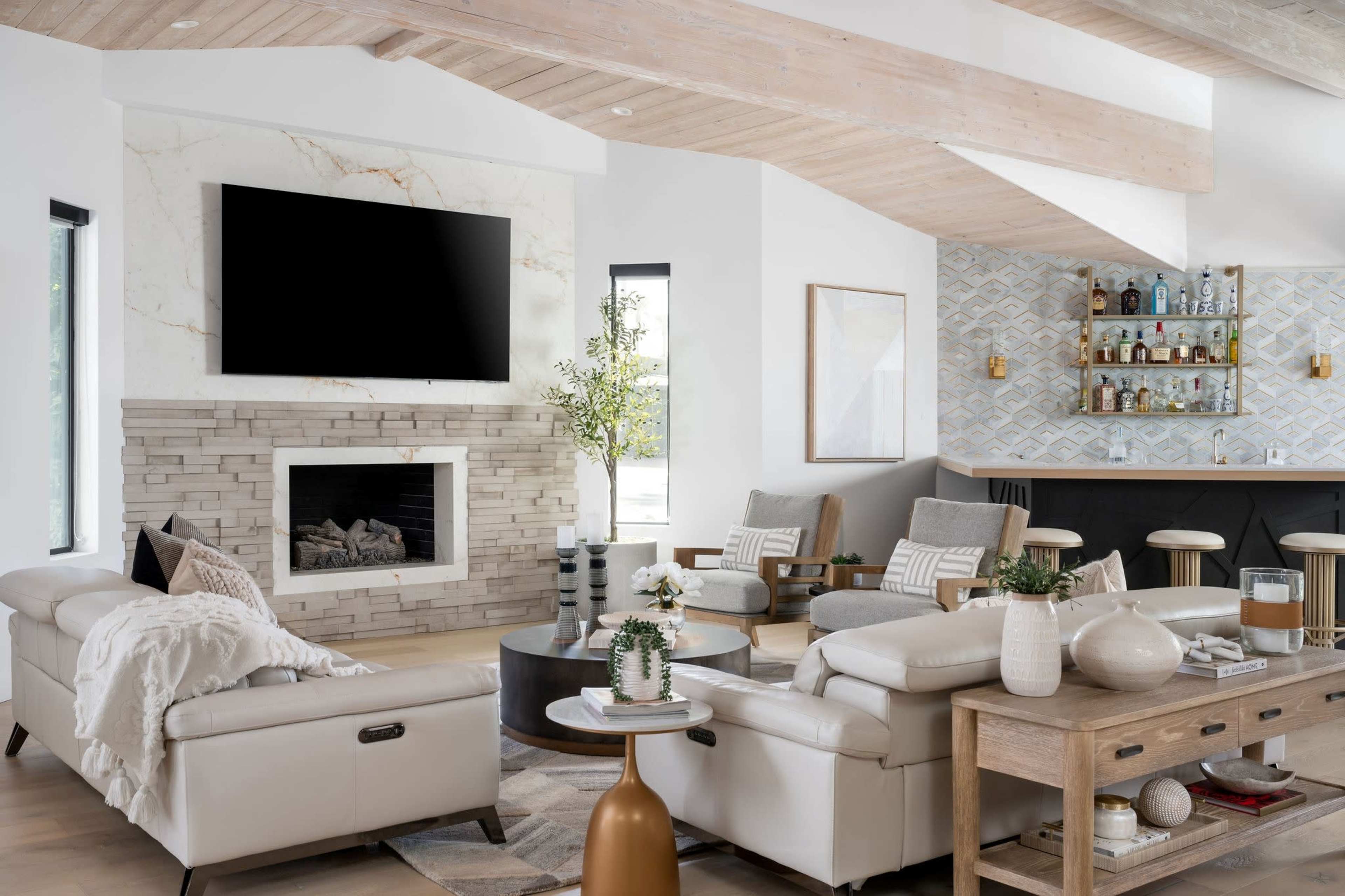 The image shows a modern living room with a stone fireplace, a large wall-mounted TV, light-colored furniture, and a bar area featuring decorative bottles.