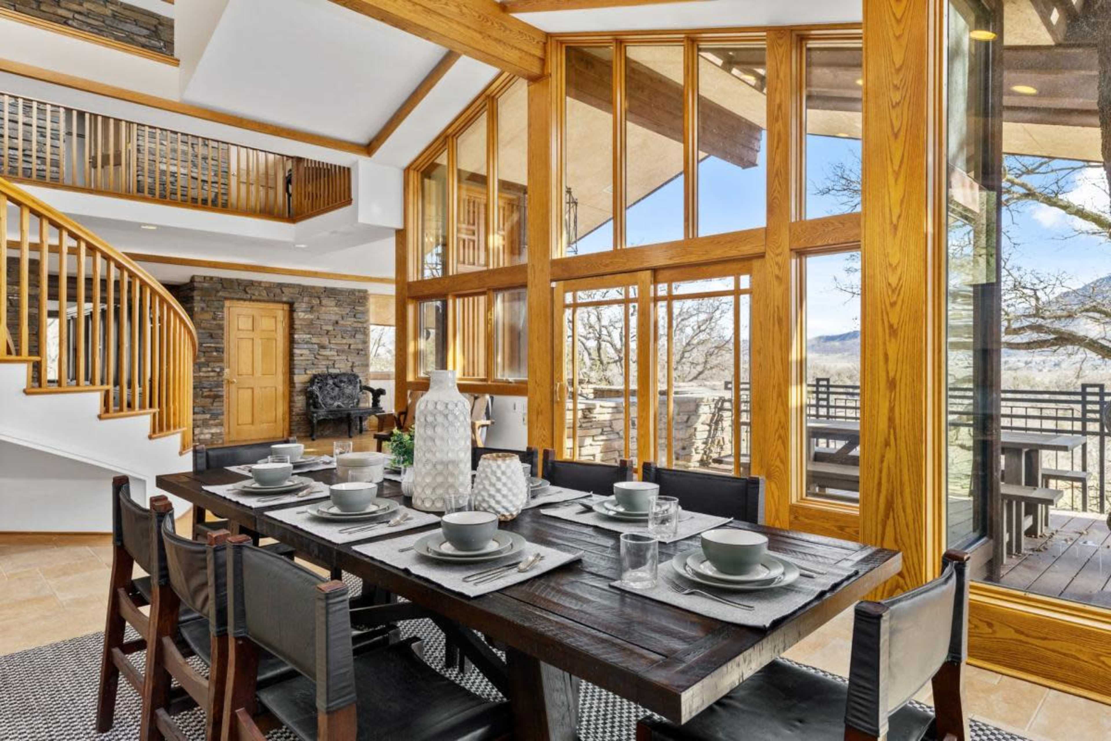 A modern dining area features a large wooden table set with dishes, surrounded by windows that provide a view of the outdoors.