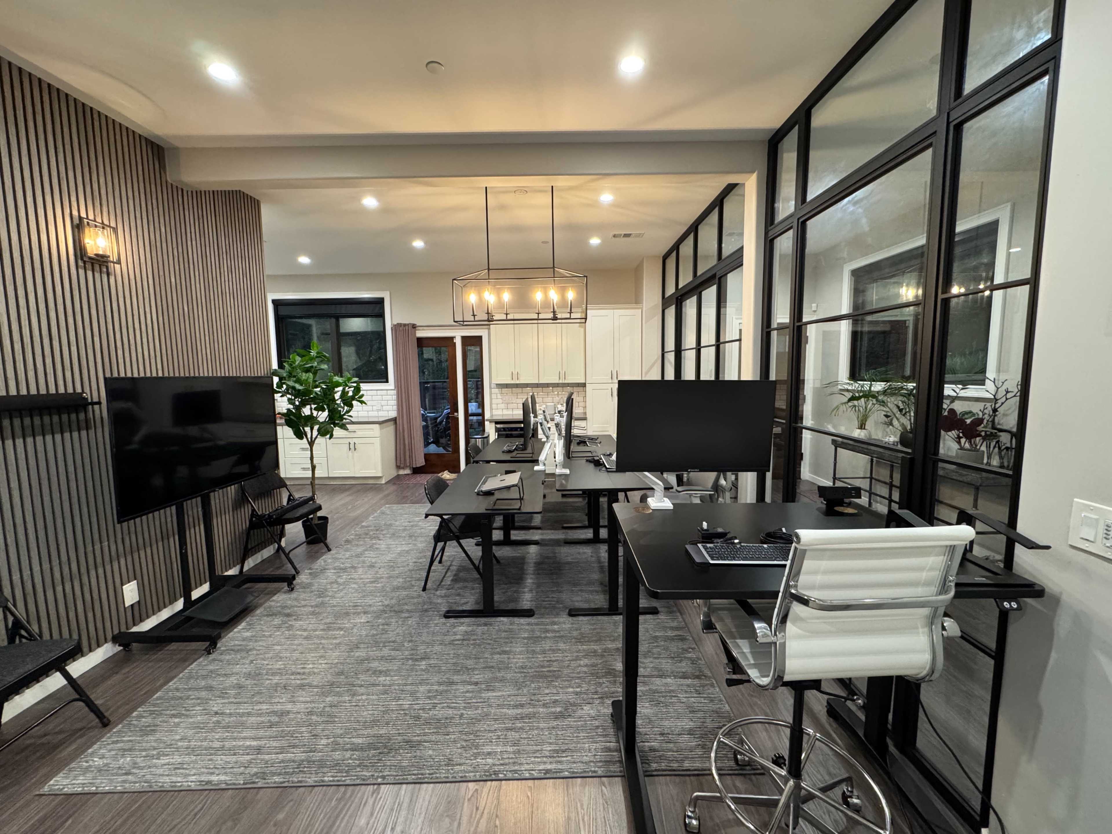 The image shows a modern office space with multiple workstations, large windows, and a central dining area featuring a chandelier.