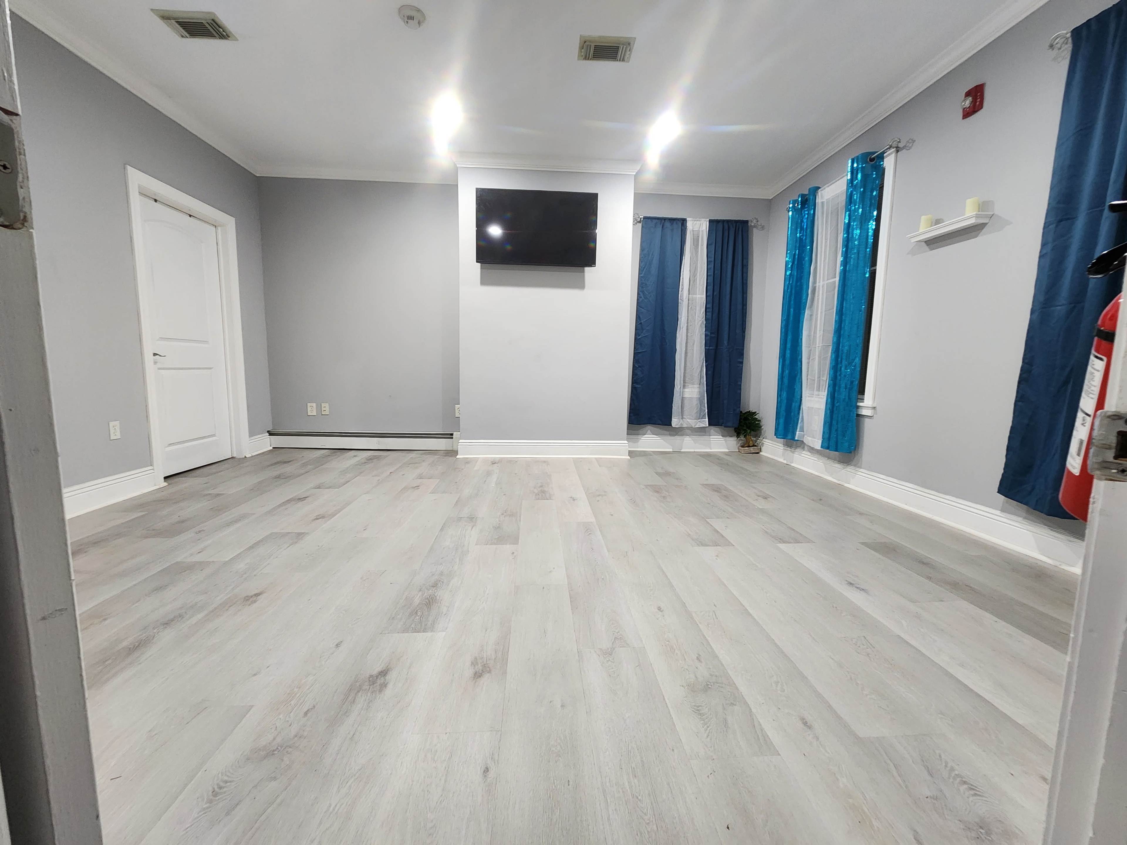 An empty room with light gray walls, a flat-screen TV mounted on the wall, and a mix of blue and white curtains at the windows.