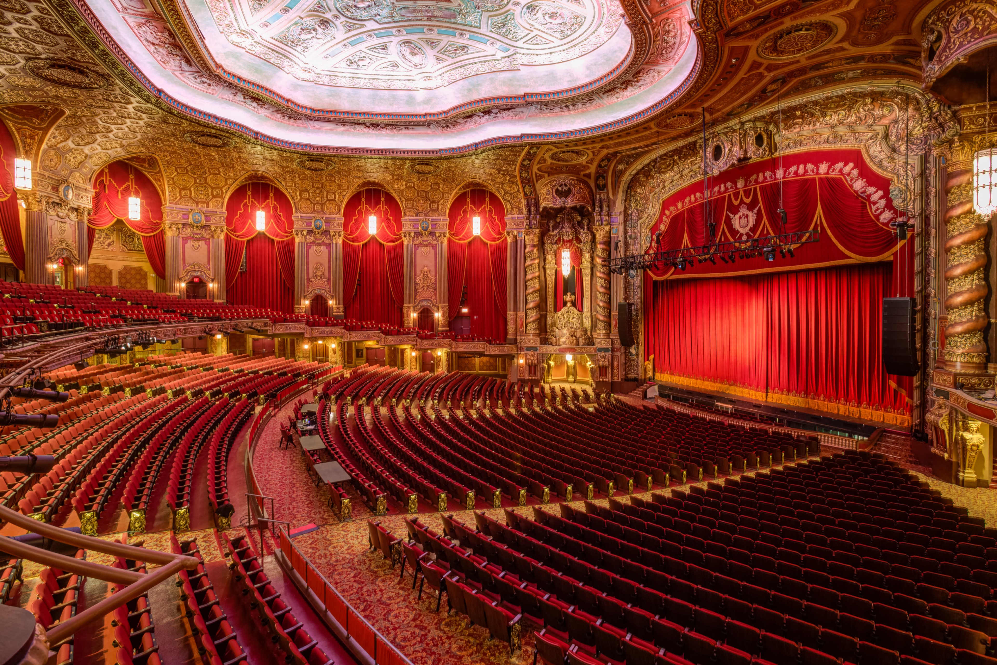 The image shows a grand theater interior with ornate decorations, red velvet curtains, and rows of empty seats facing the stage.