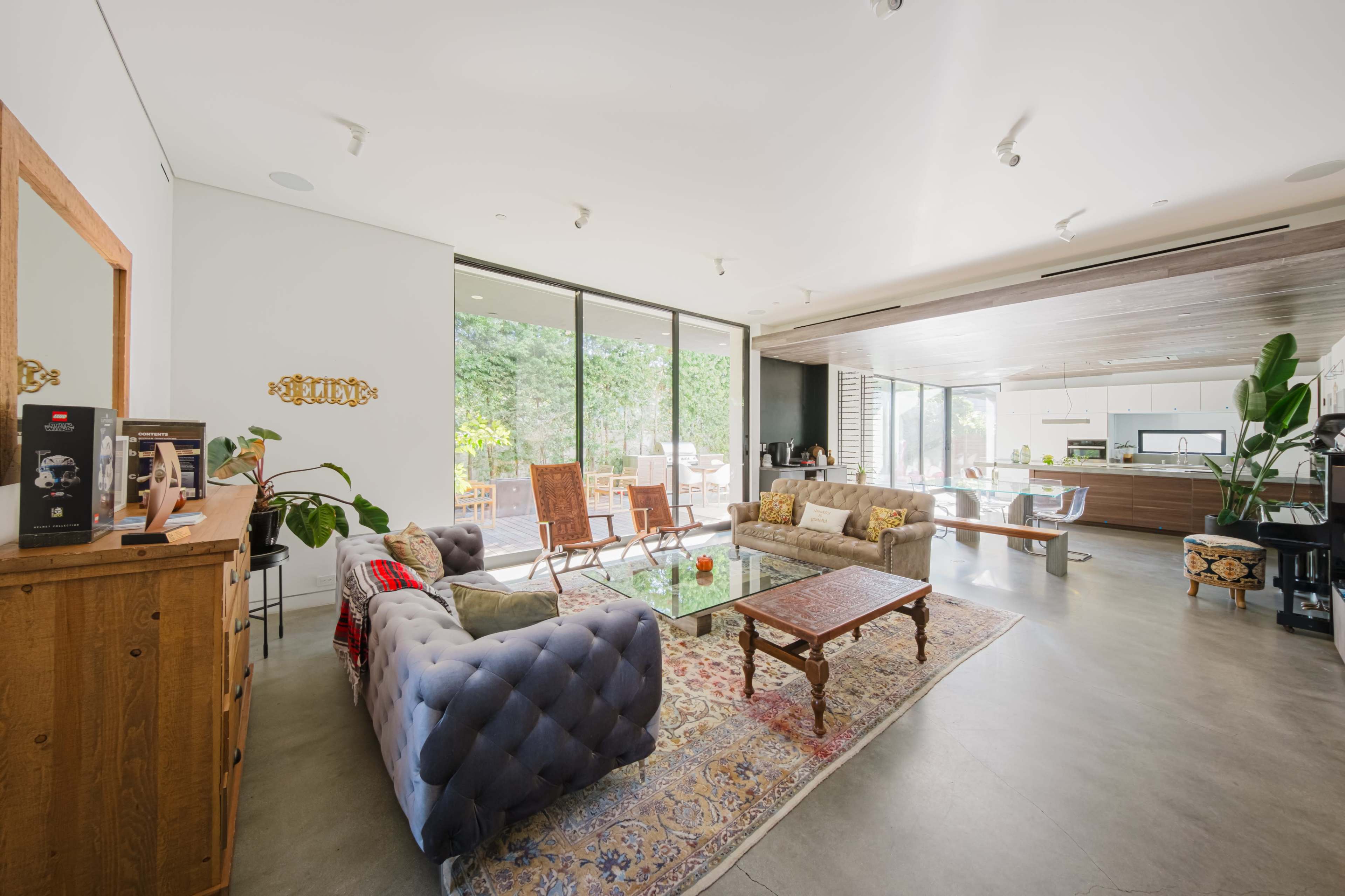 The image shows a modern living room with a large glass coffee table, a sectional sofa, and a wooden side table, featuring large windows that open to an outdoor area.