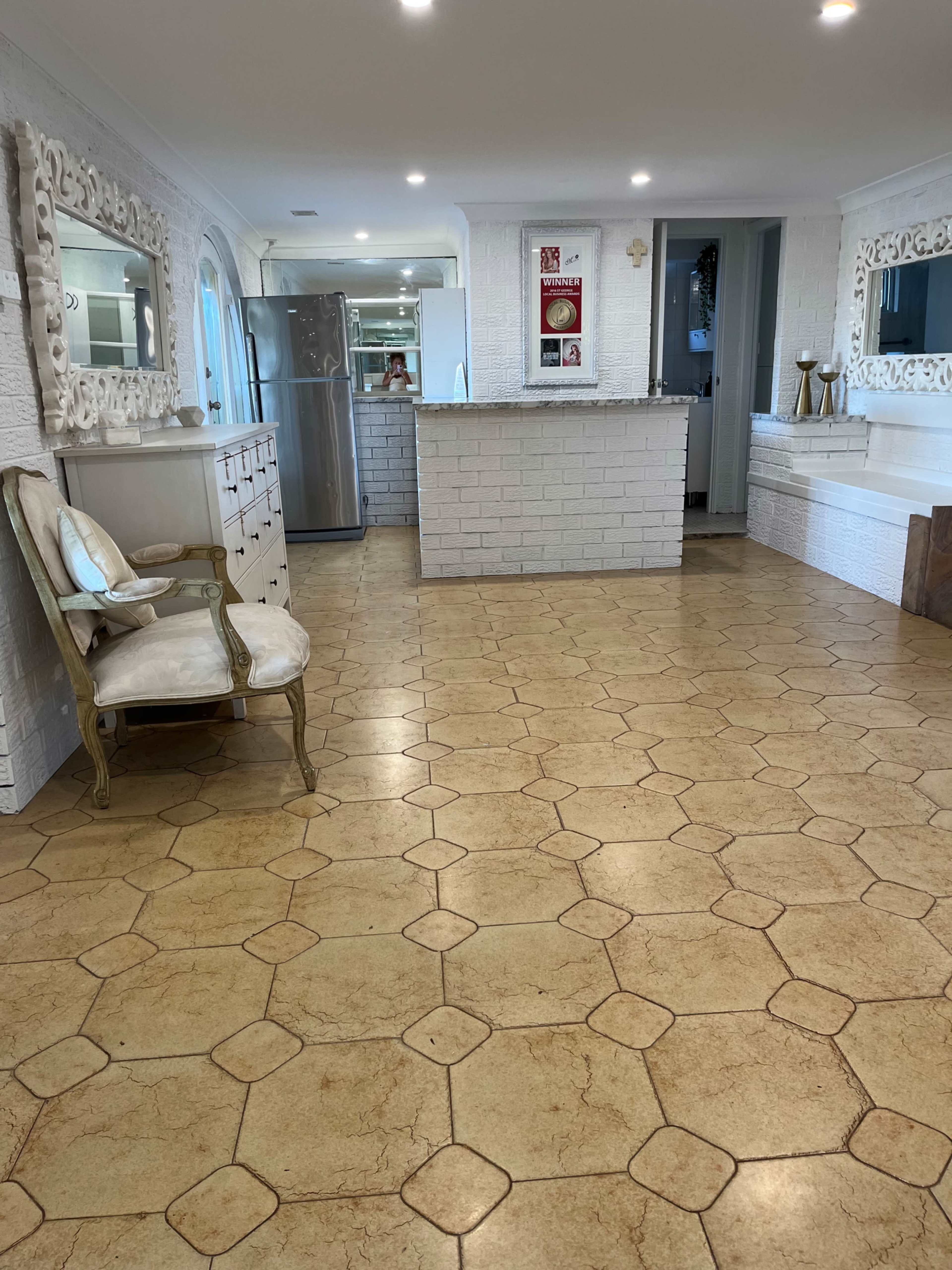 The image shows a spacious kitchen with light-colored hexagonal floor tiles, a white brick wall, and a bar-style counter behind a seating area with a decorative chair.