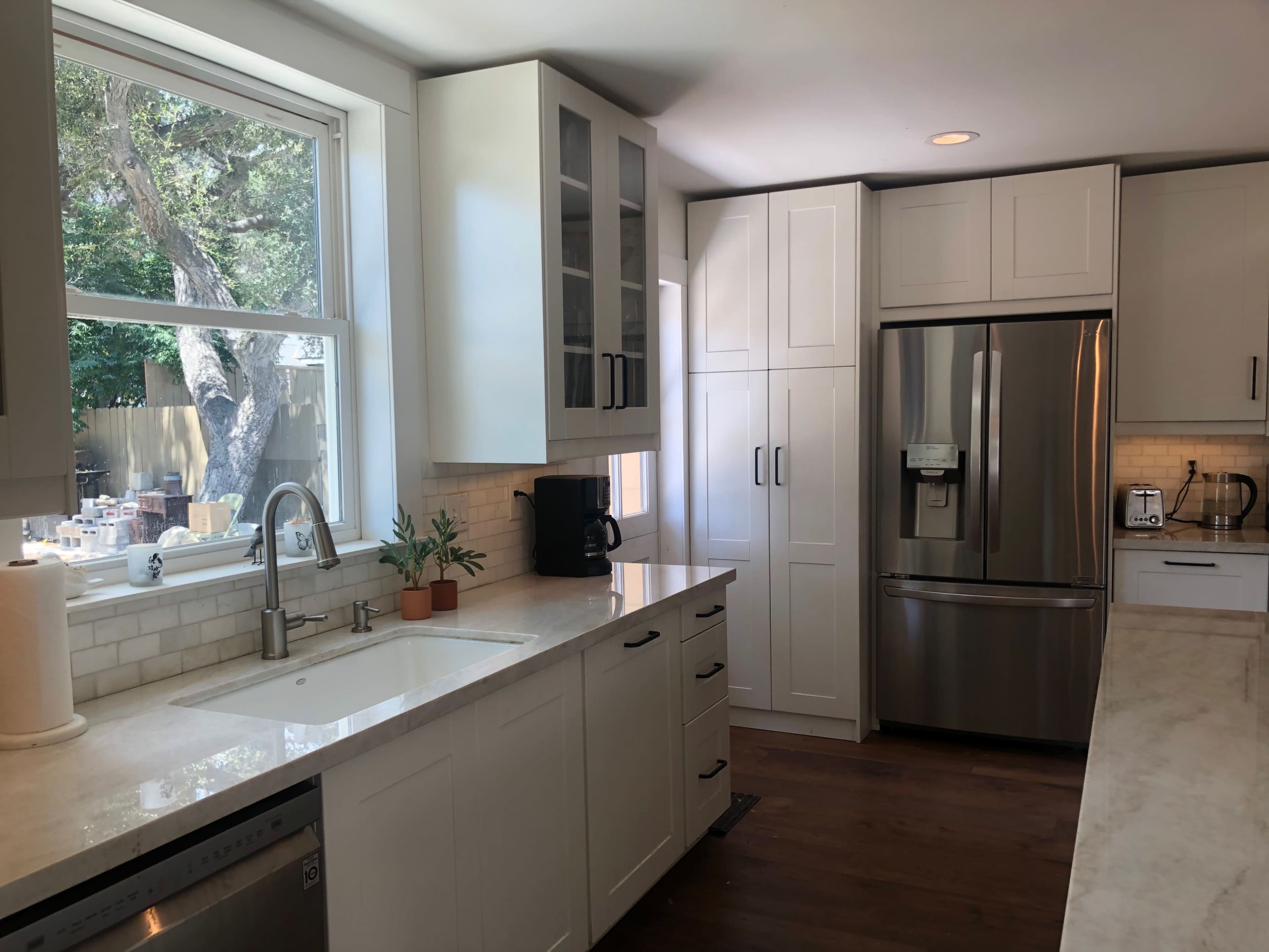 A modern kitchen featuring white cabinets, a stainless steel refrigerator, and a large window overlooking a tree.