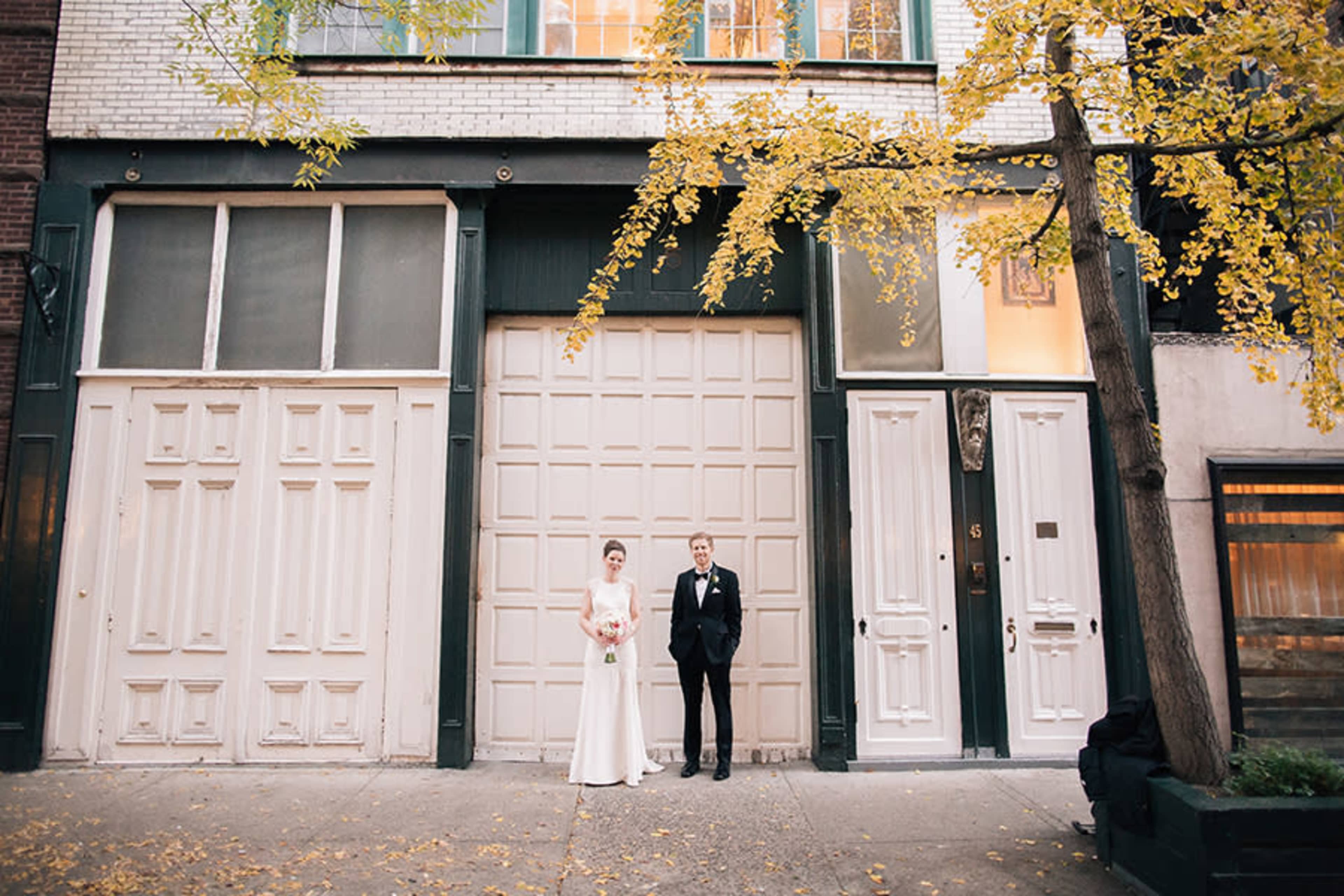 A bride and groom stand in front of two residential doors, surrounded by yellow autumn leaves.