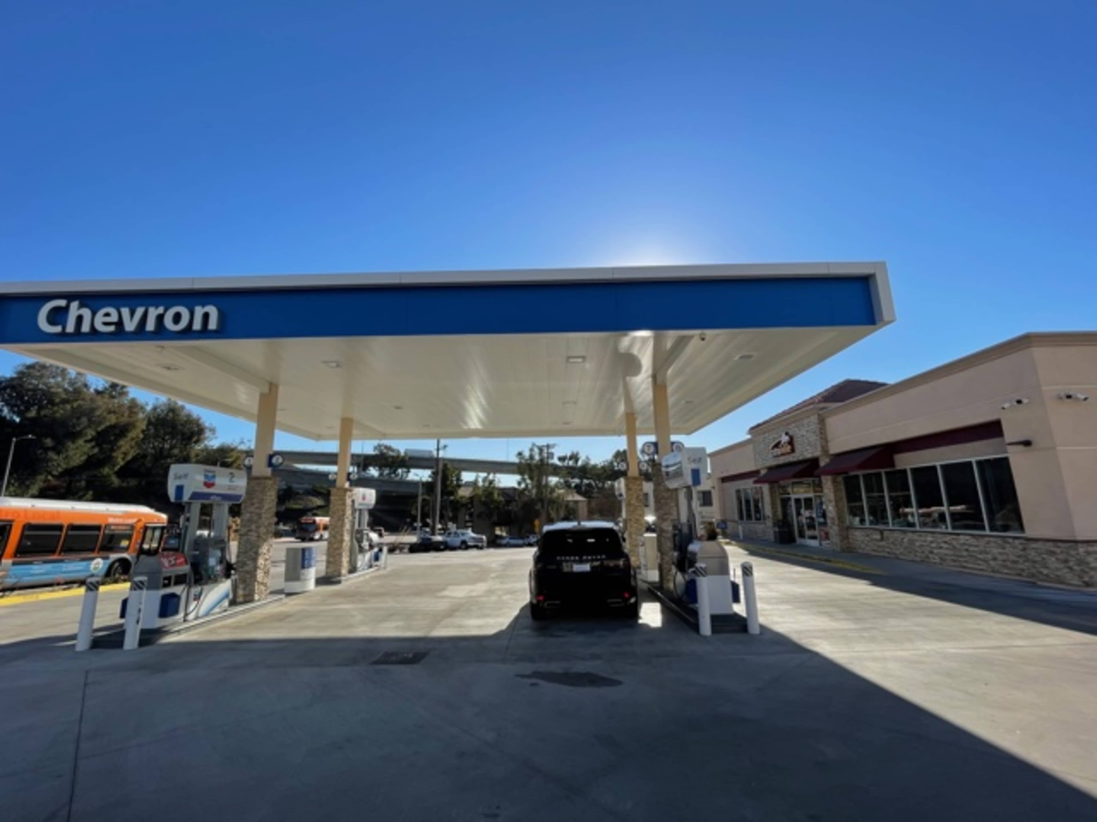 A Chevron gas station with a clear sky and a nearby building is shown, featuring a black vehicle parked at one of the gas pumps.