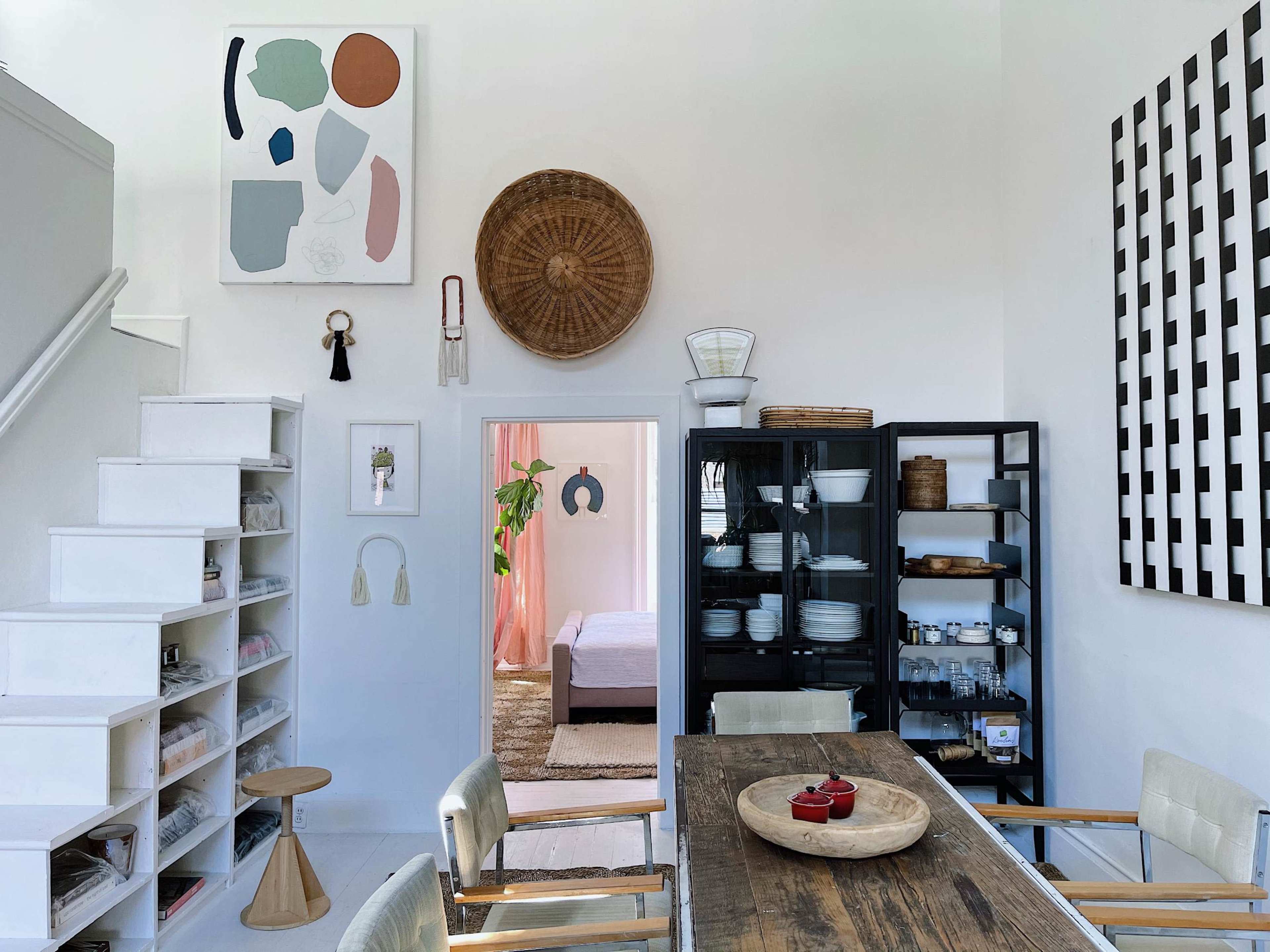 A bright dining area features a wooden table, chairs, and various storage solutions, with art on the walls and an open space leading to a bedroom.