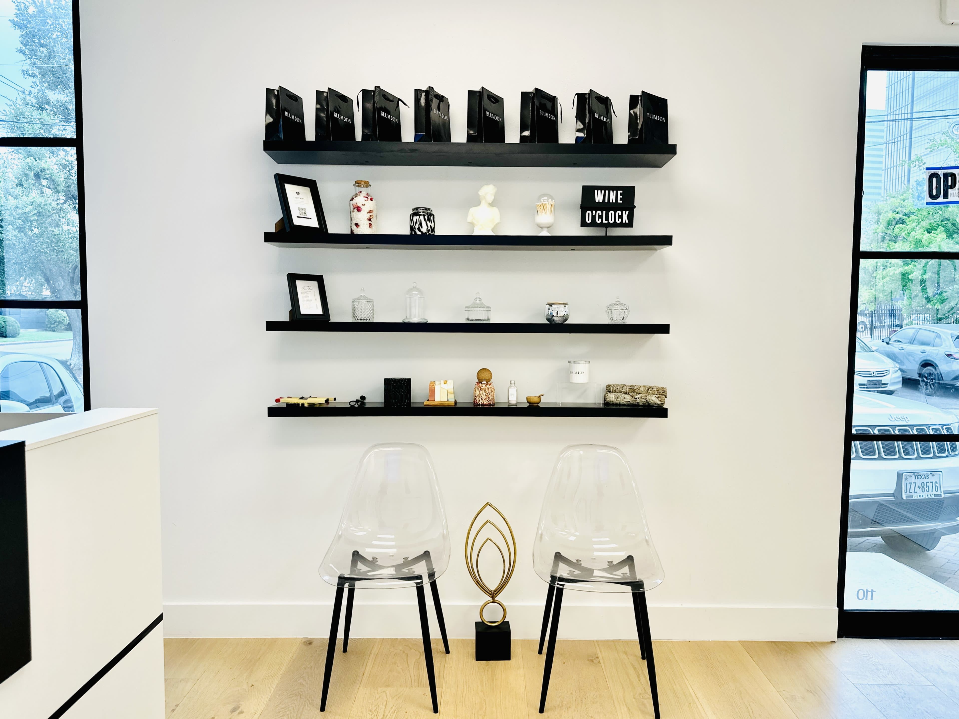 A minimalist interior features a wall shelf displaying various decorative items and trophies above two clear plastic chairs.