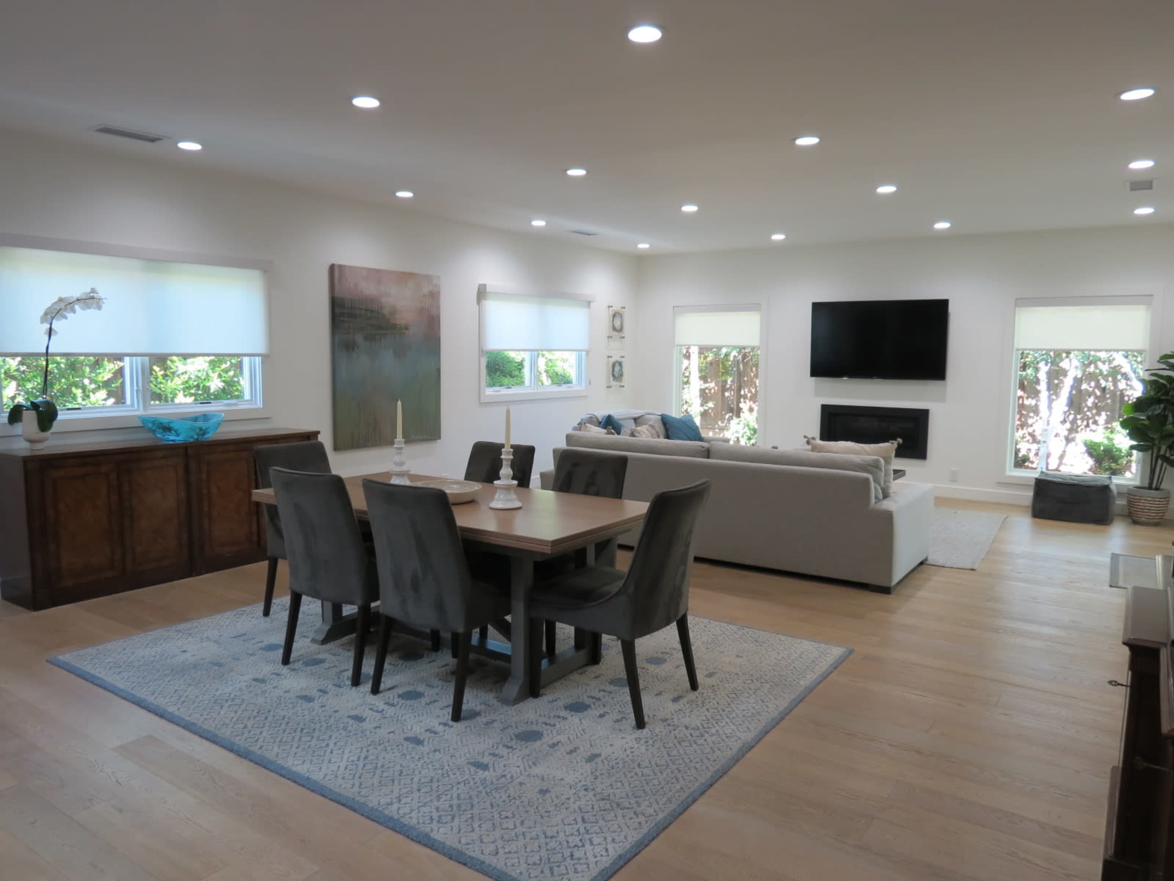 A bright and spacious living area features a dining table, gray chairs, a large sofa, and a television mounted on the wall, with windows allowing natural light to fill the room.