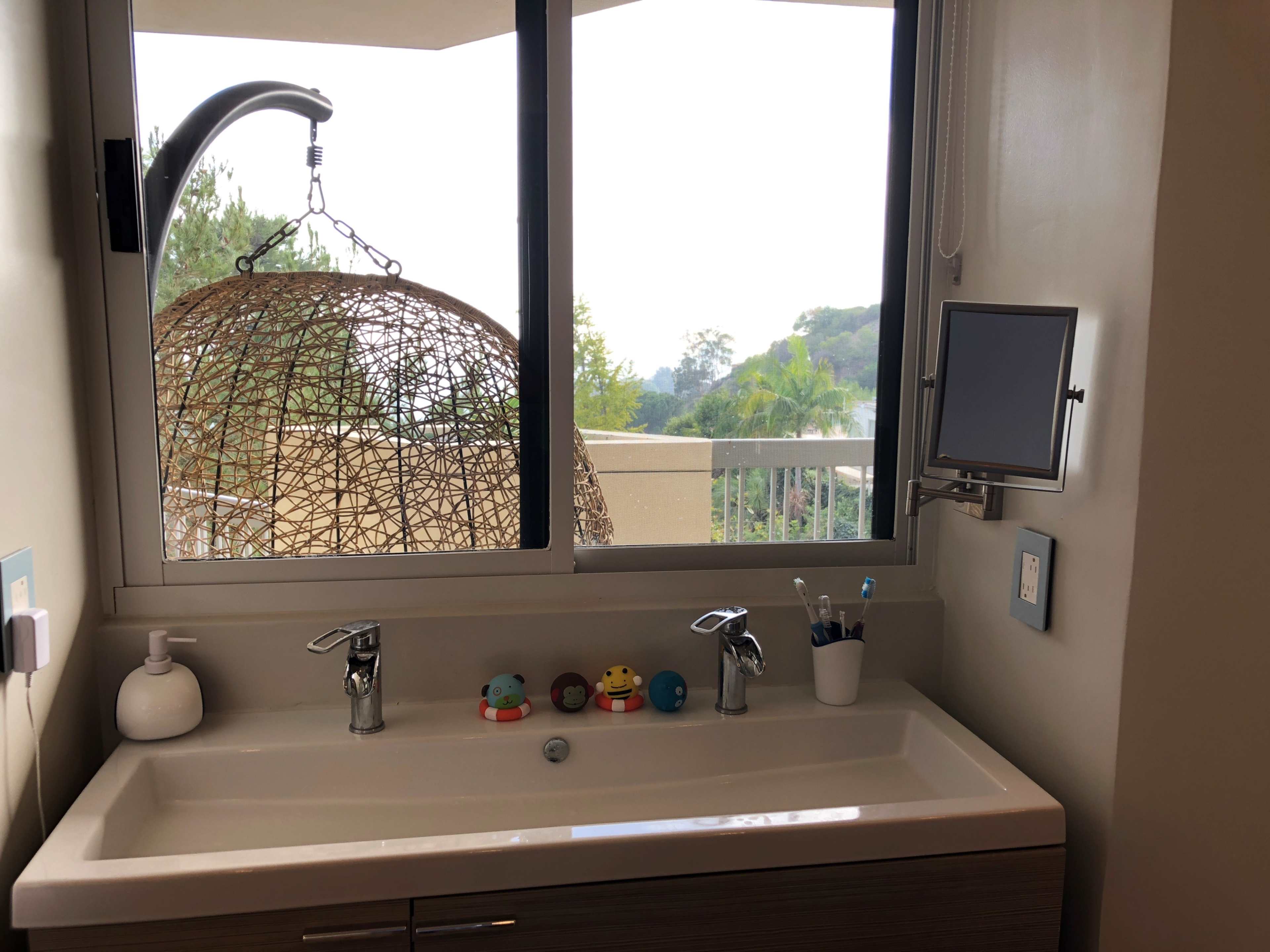 A bathroom sink with a decorative mirror, several small rubber ducks, and a window overlooking a balcony and greenery.