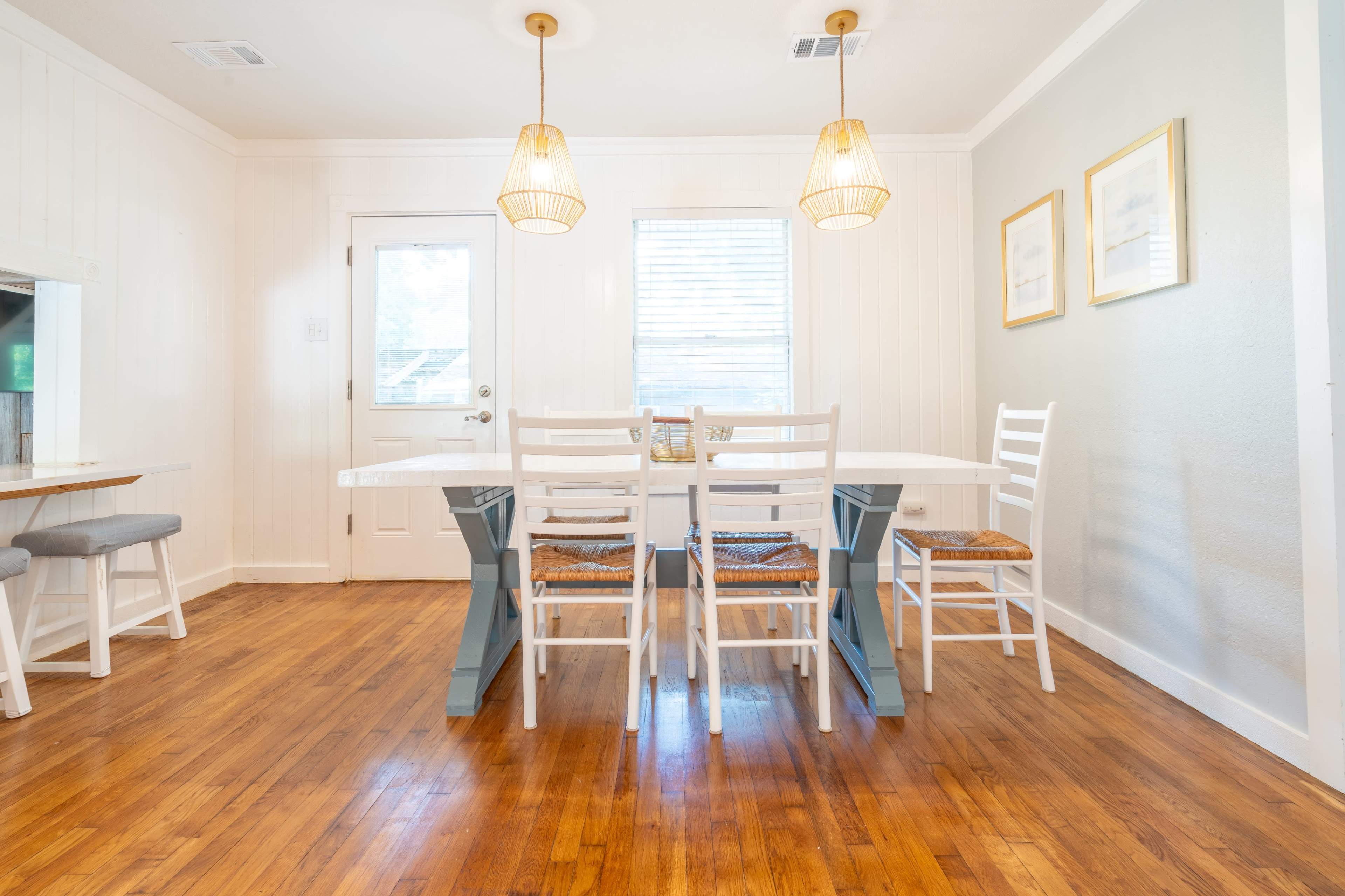 A bright dining area features a long white table surrounded by six white chairs, with two pendant lights overhead.