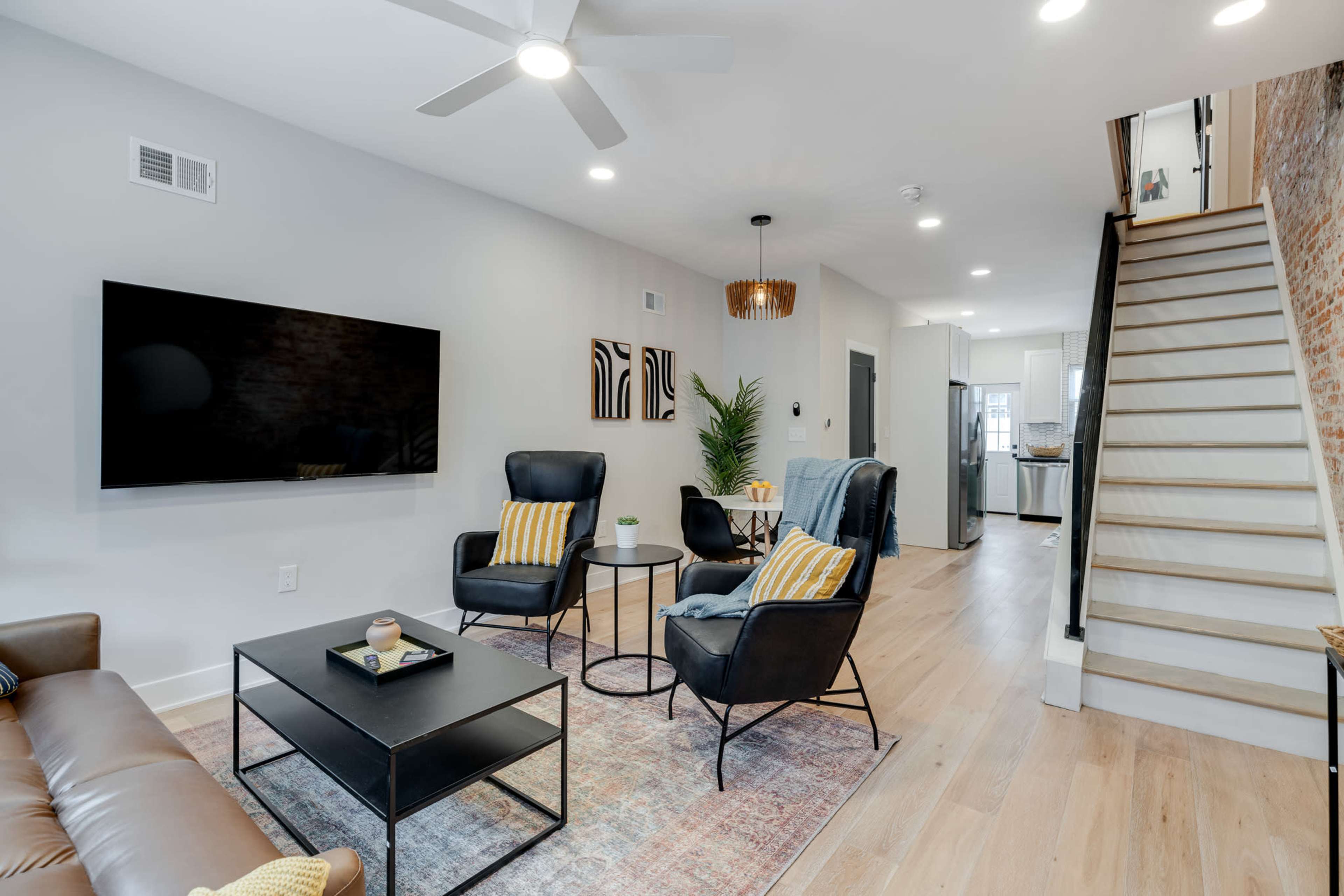 A modern living room features a TV mounted on the wall, two black chairs with yellow-striped cushions, a coffee table, and an open staircase leading to the upper floor.