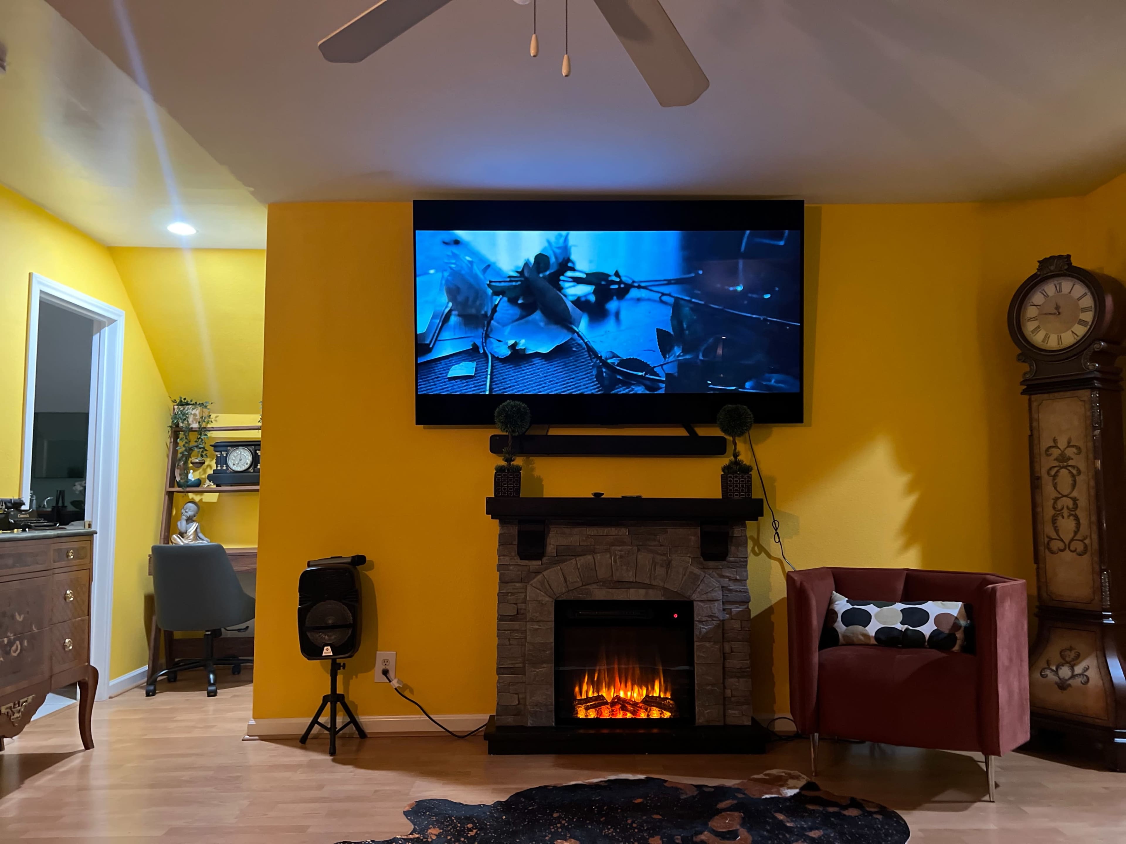 A large television displaying a movie scene is mounted on a yellow wall above a stone fireplace, with a red armchair and a desk visible in the background.