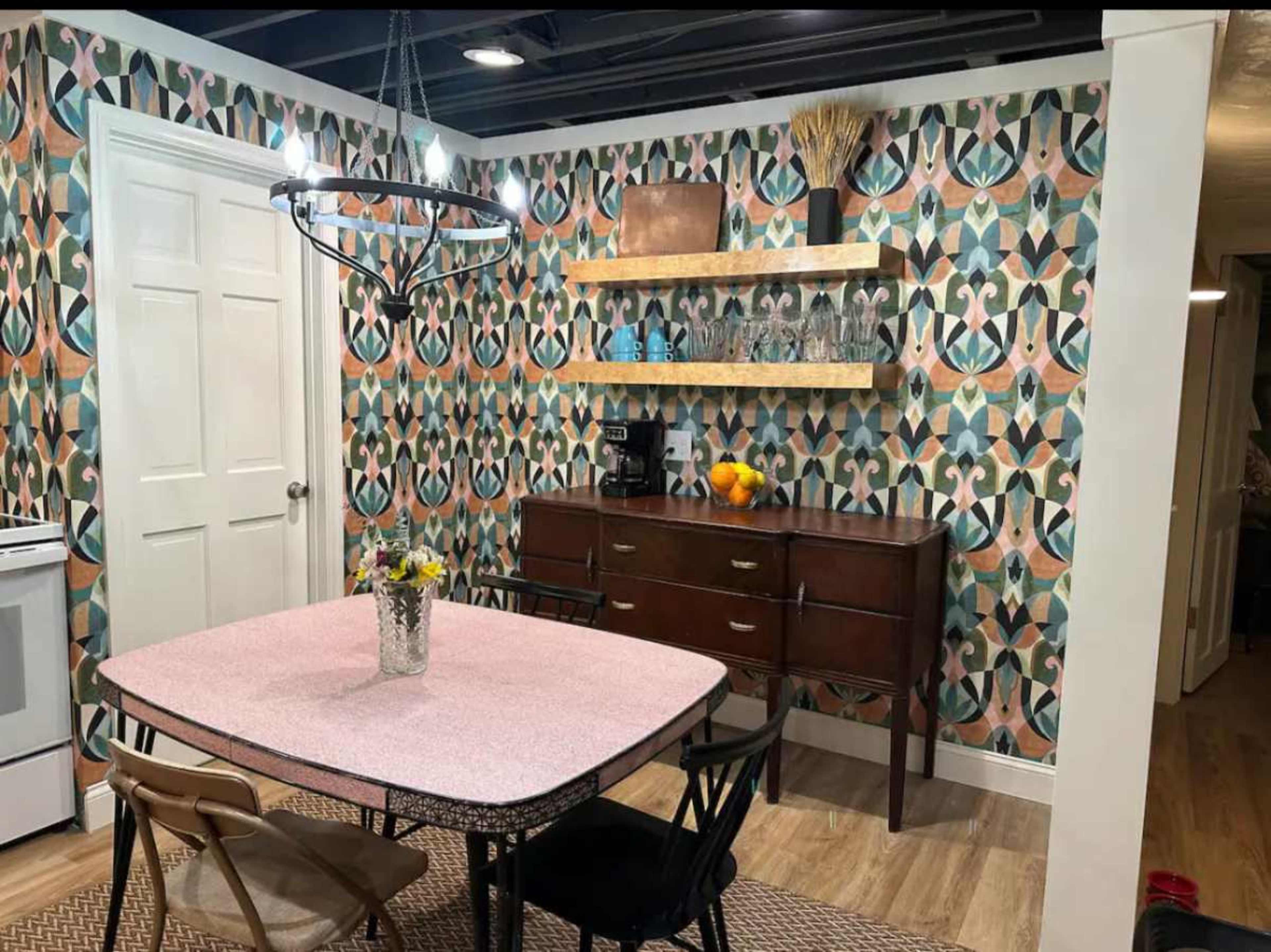 A kitchen features a pink table with black chairs, a wooden sideboard, and patterned wallpaper adorned with geometric designs.