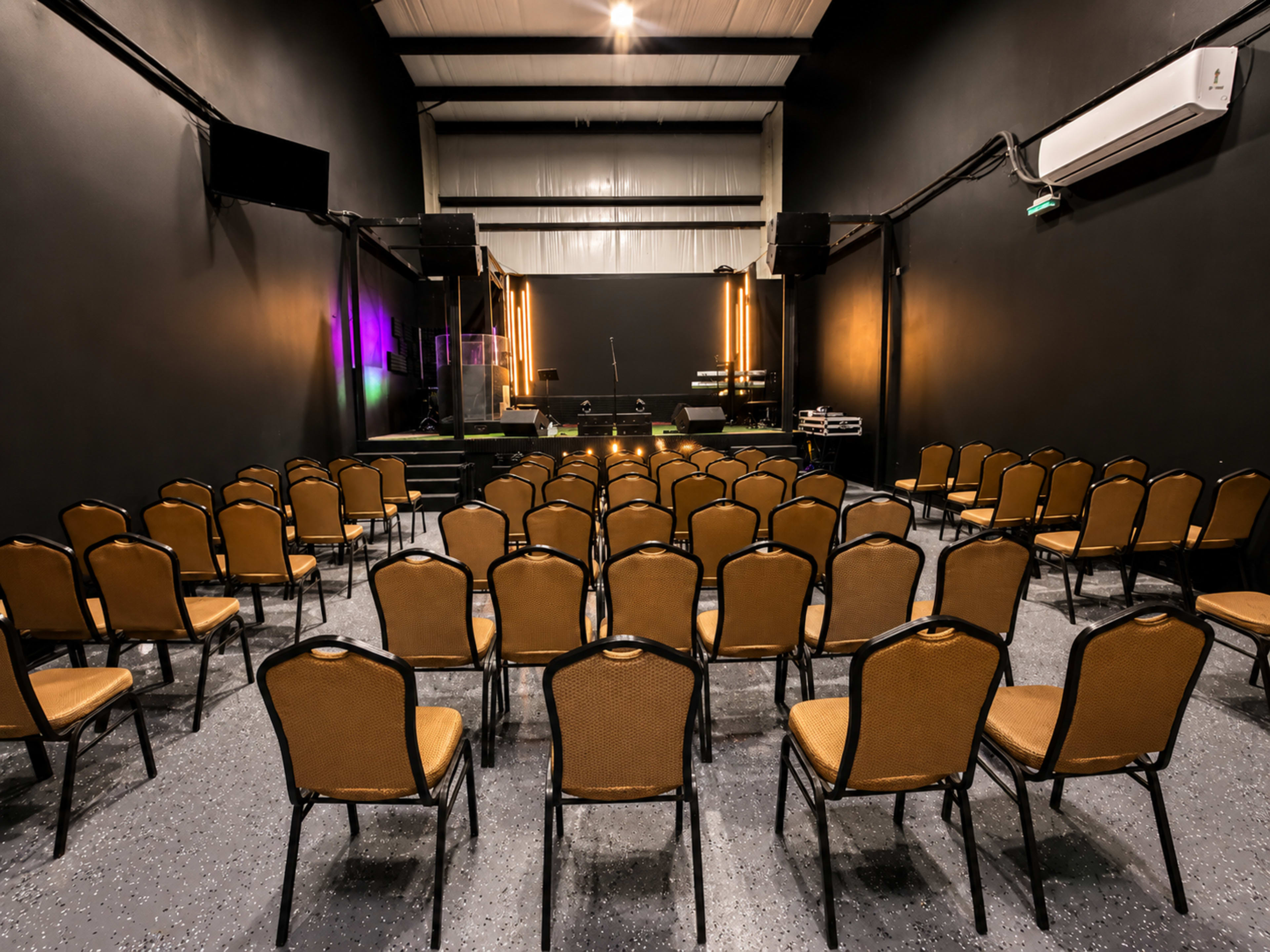 The image shows an empty music venue with rows of chairs facing a stage, set against black walls and dim lighting.