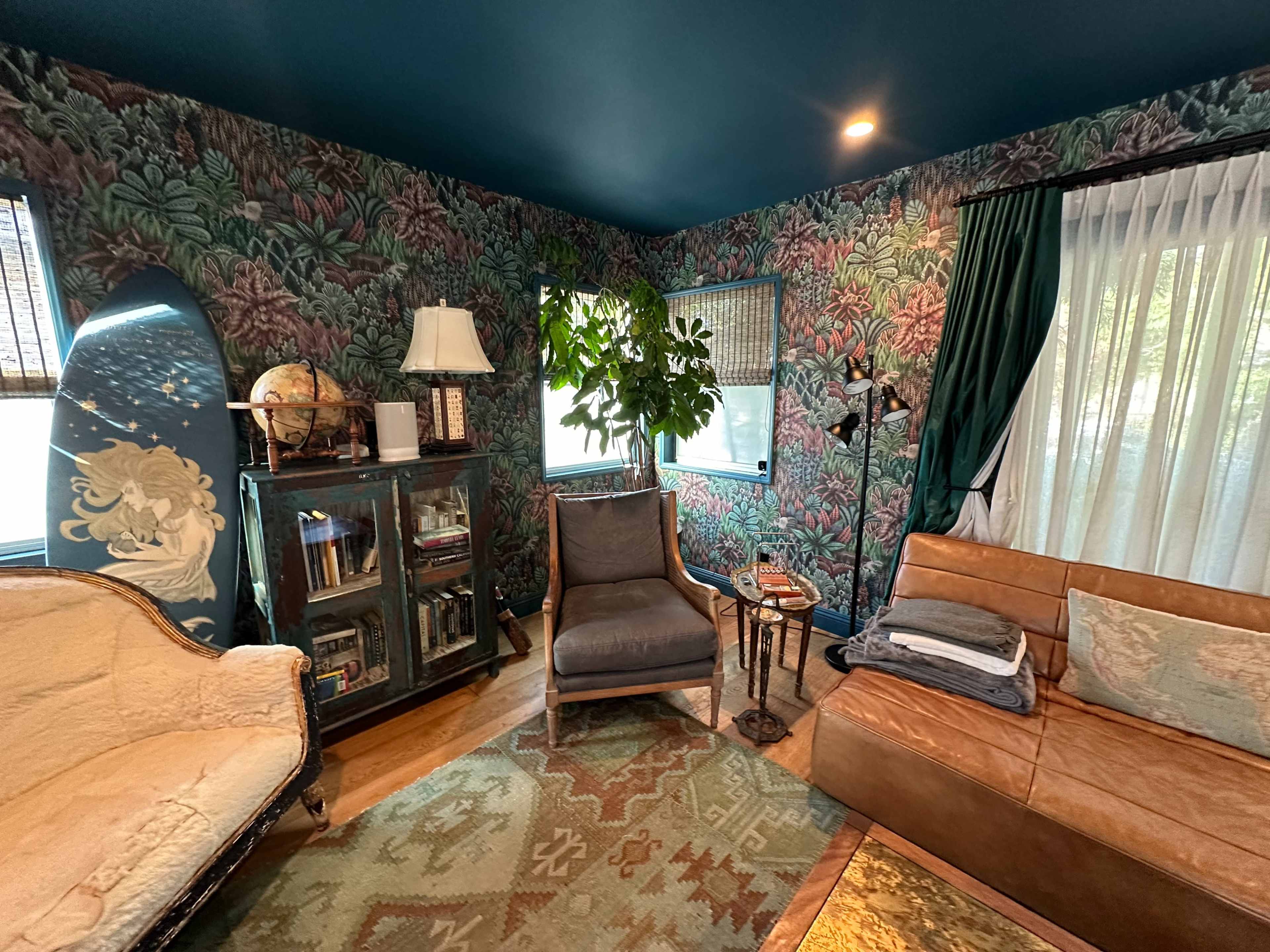 The room features a vintage armchair, a leather sofa, a bookshelf, and large windows framed by sheer curtains and patterned wallpaper.