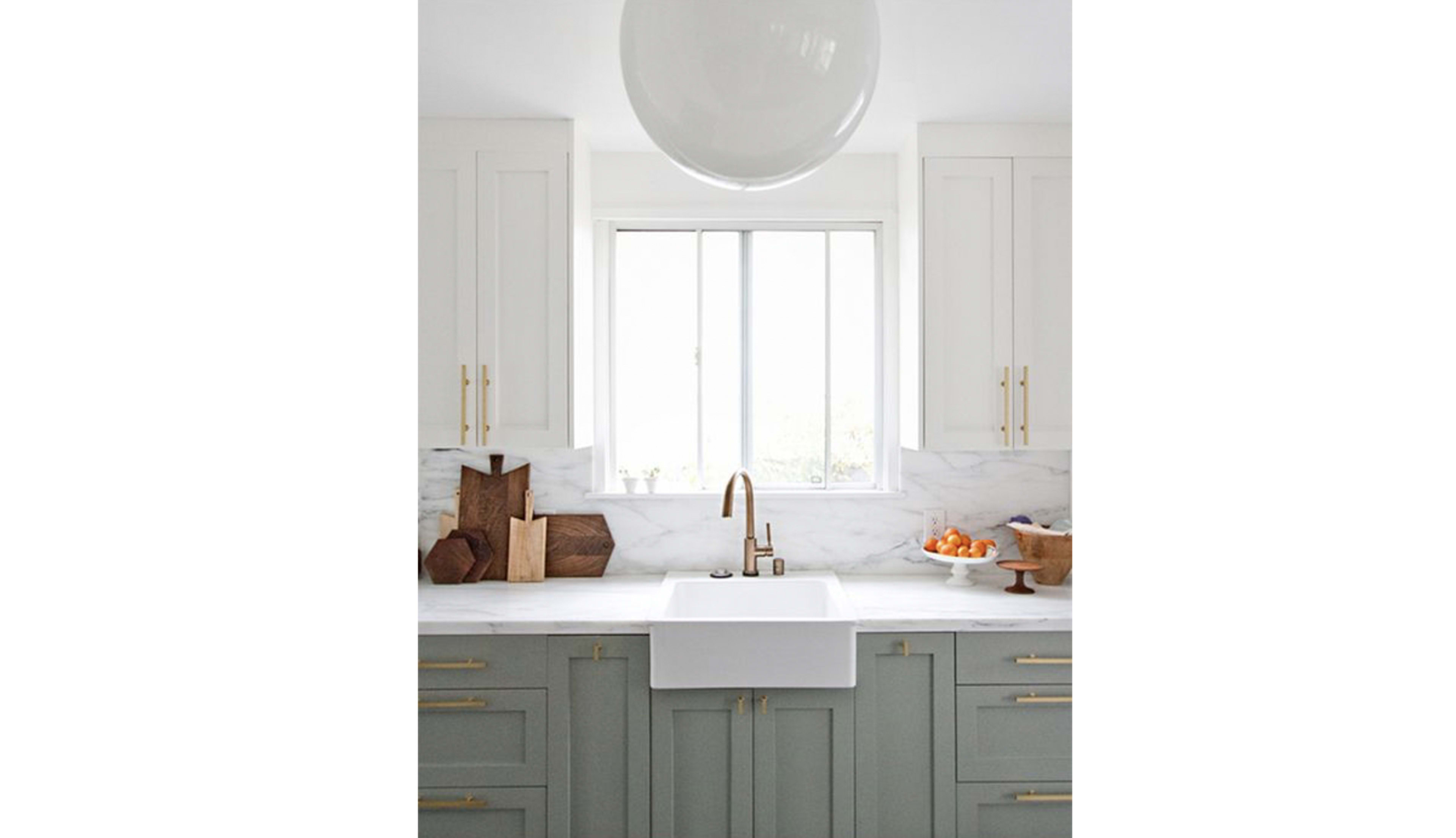 A modern kitchen with a farmhouse sink, light green cabinetry, marble countertops, and a window above the sink.