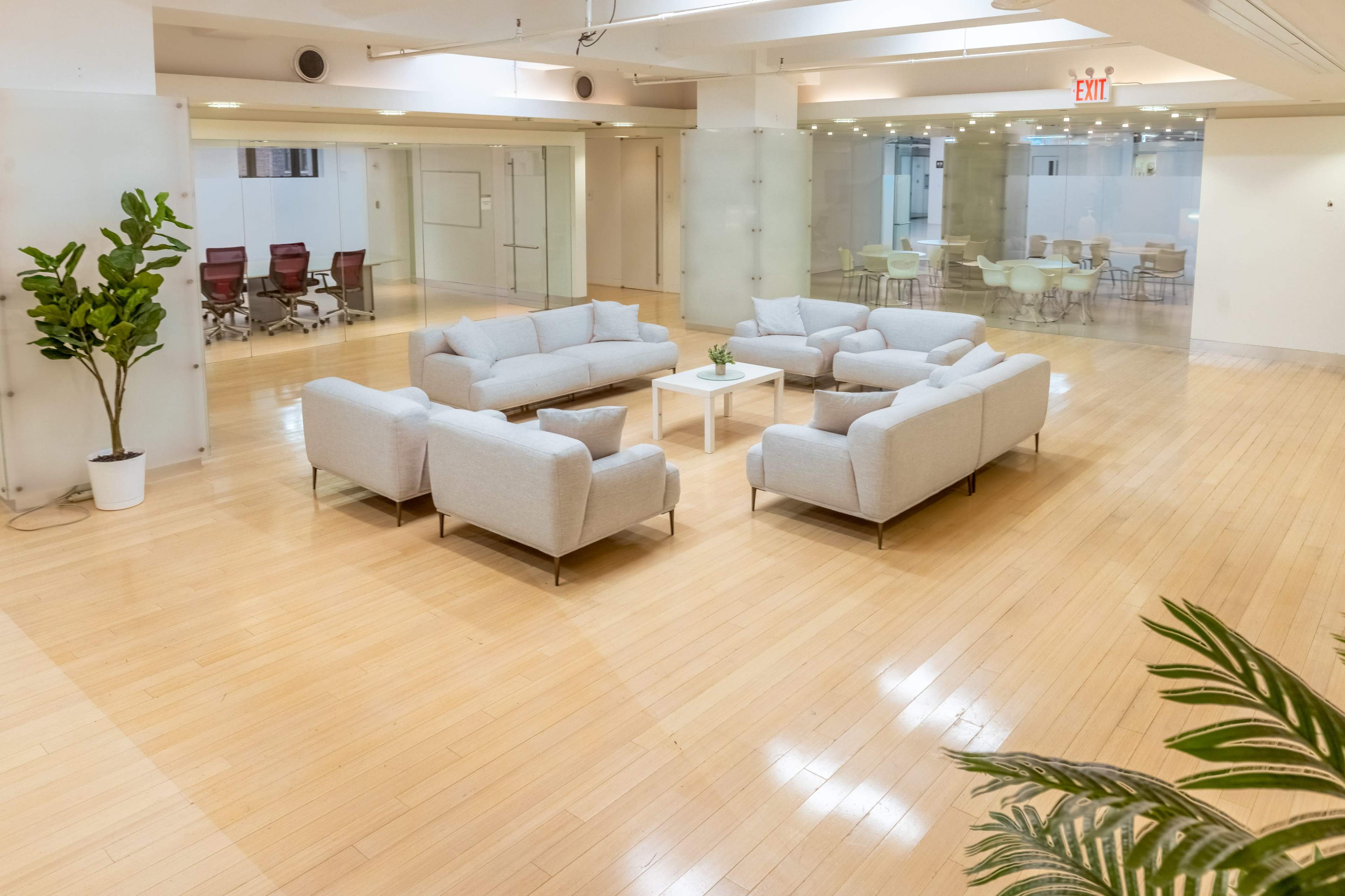 The image shows a modern office space featuring two sets of light-colored sofas arranged around a central table, with glass-walled meeting rooms in the background.