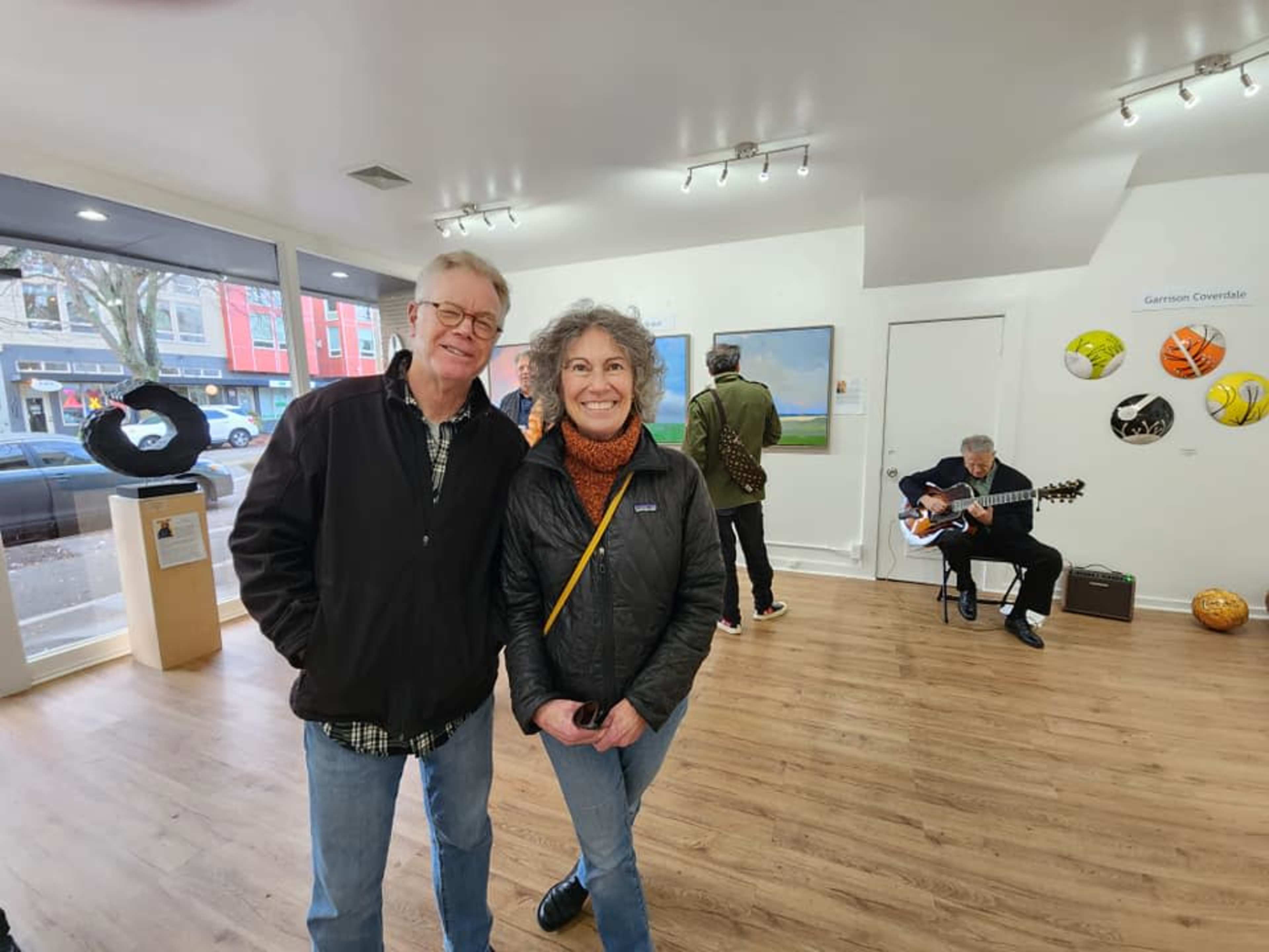 Two people stand smiling in an art gallery, while a musician plays guitar in the background.