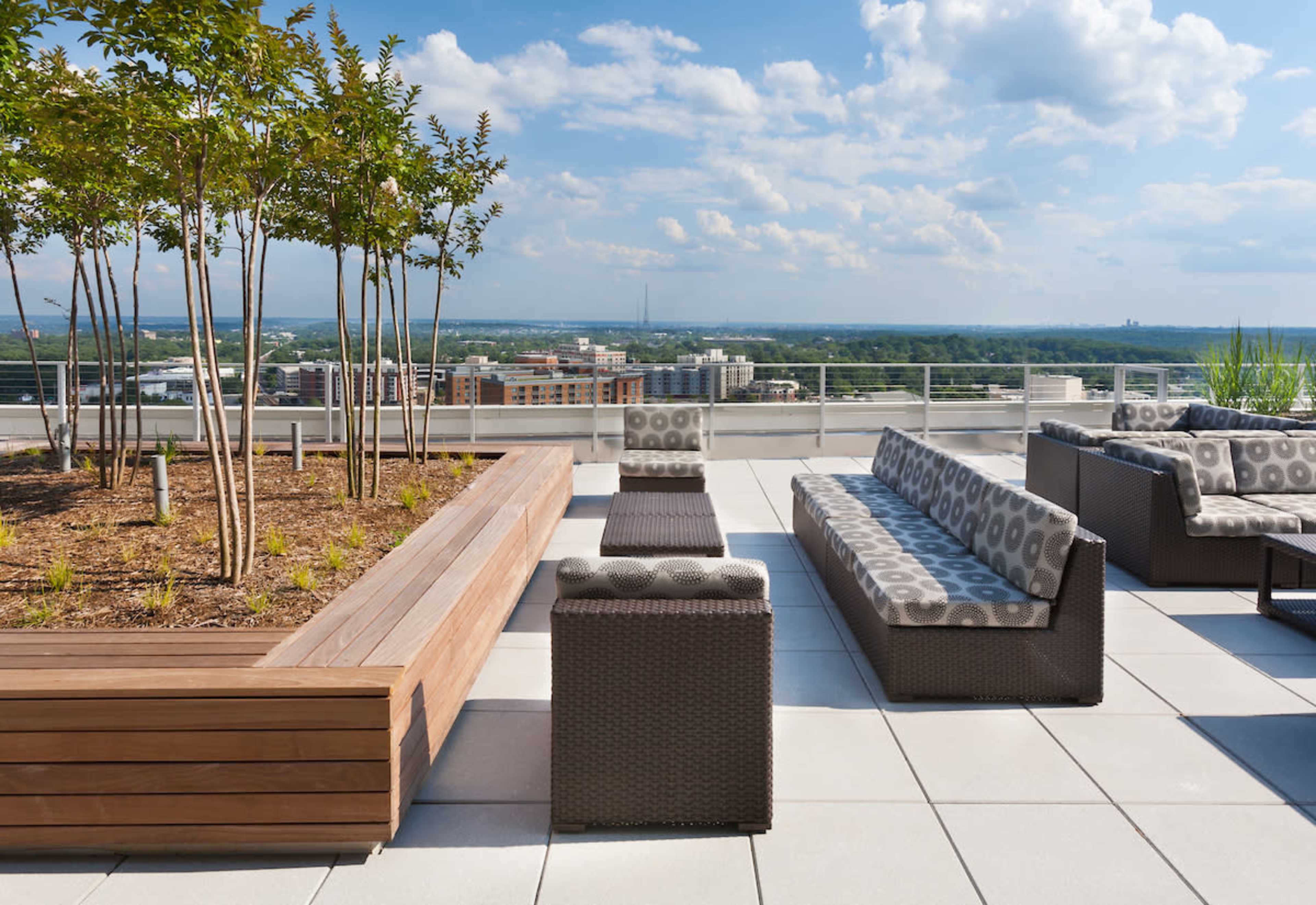 A rooftop terrace with modern seating arrangements and planters, overlooking a cityscape under a cloudy sky.
