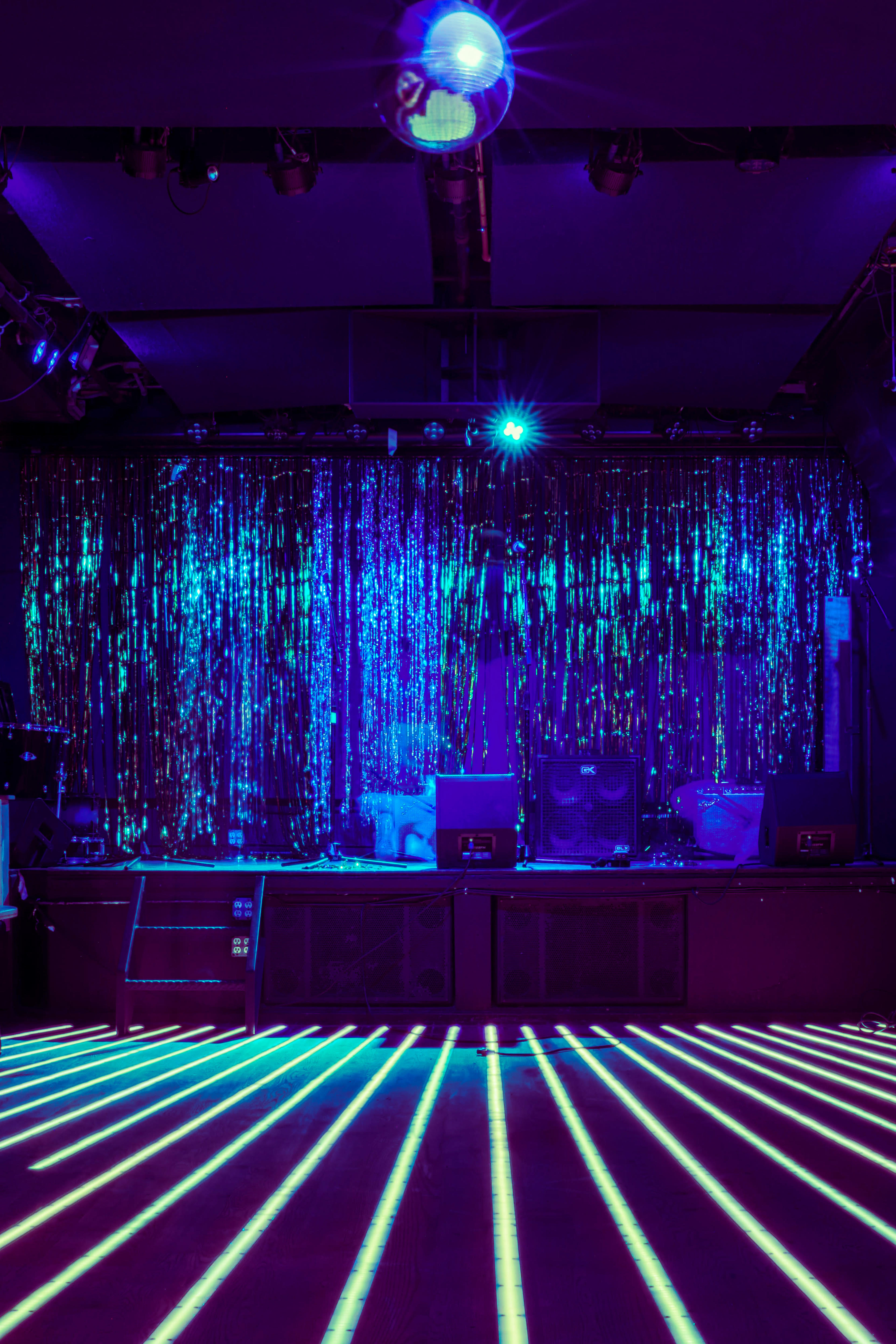 A stage is set with a shimmering curtain and colorful lights, featuring a disco ball above and illuminated floor lines leading towards it.