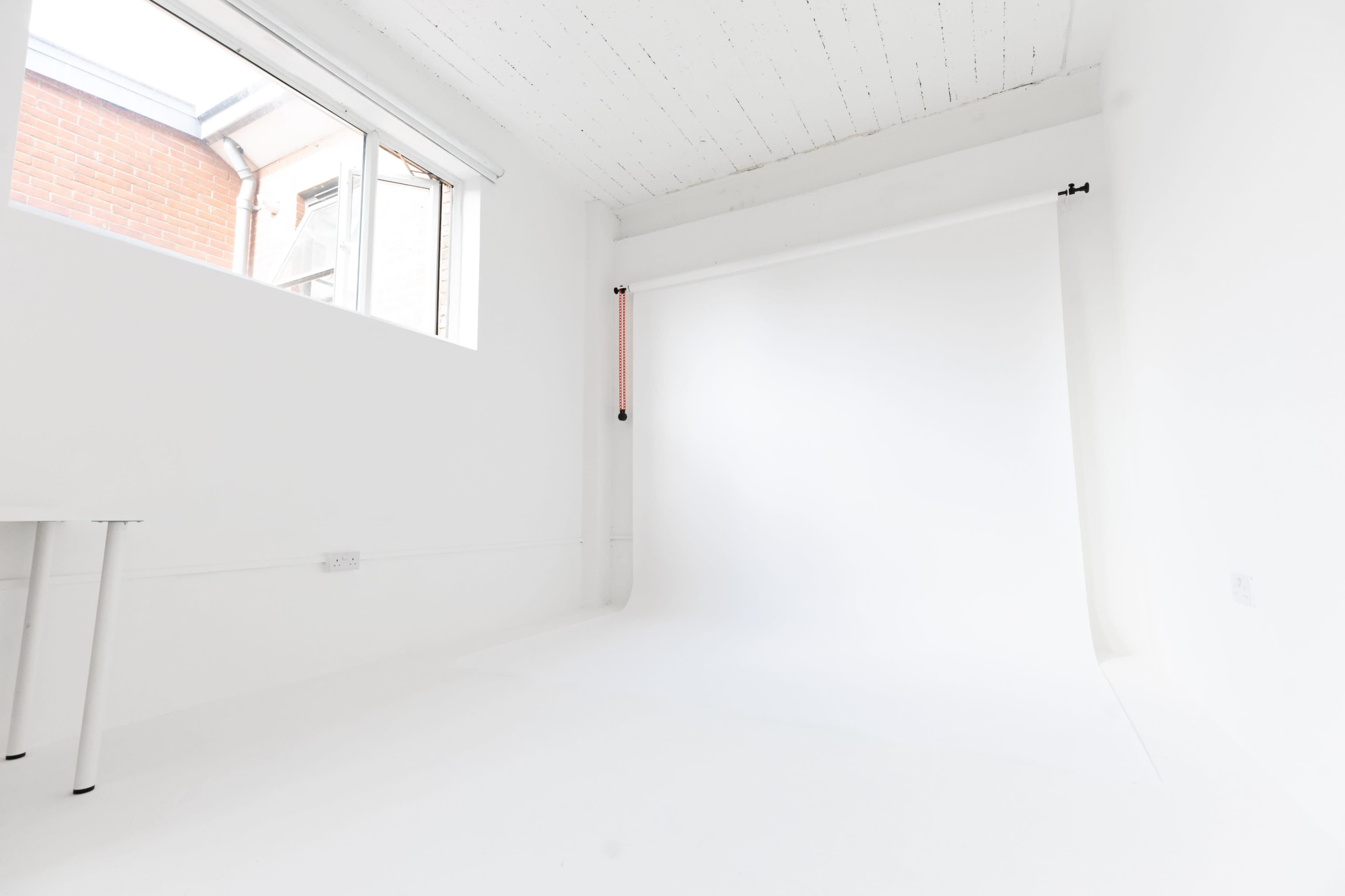 The image shows a bright, empty studio space with a white backdrop and a table in one corner.