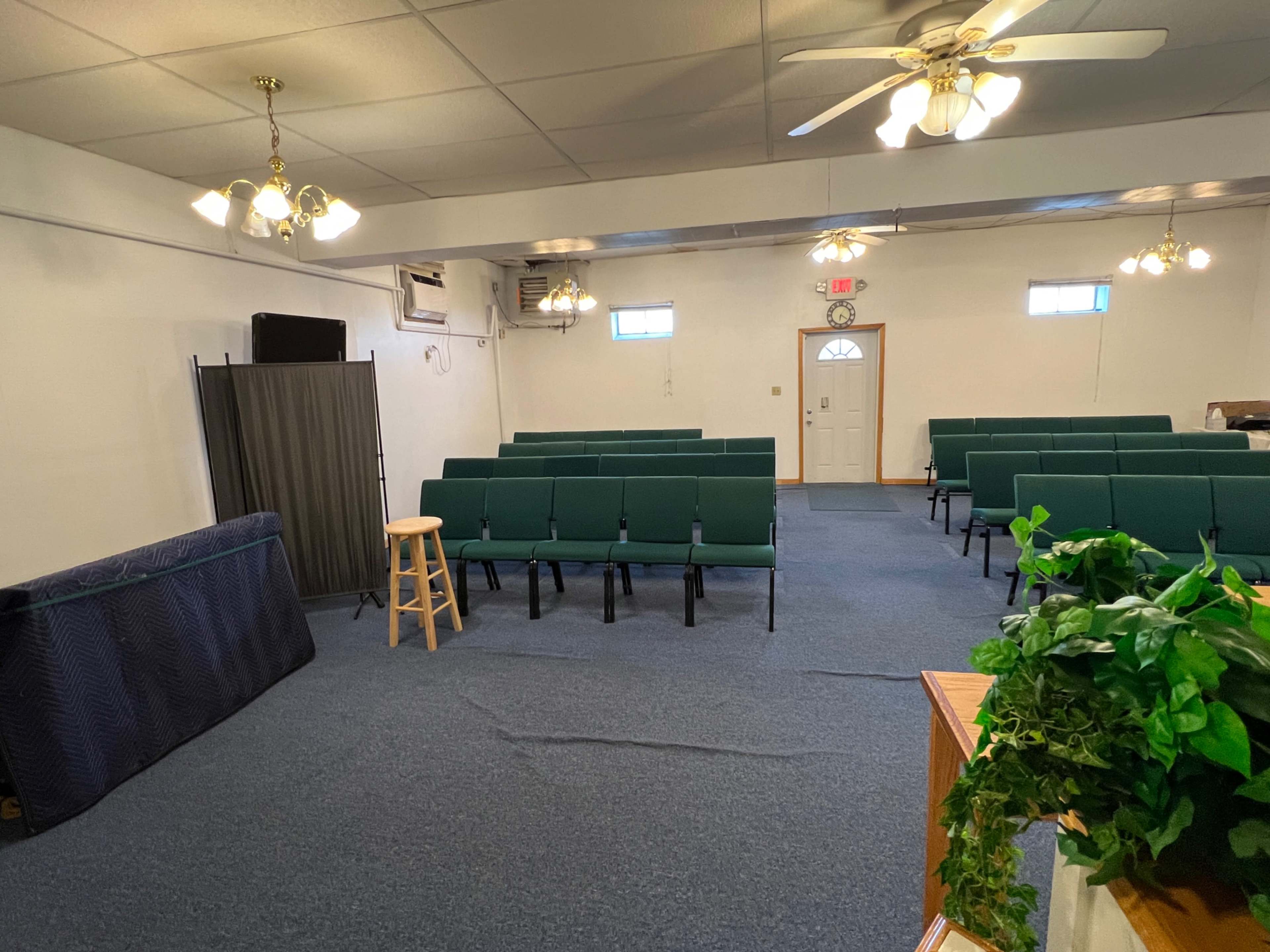 The image shows a sparsely furnished room with green chairs arranged in rows, a small stool, and a low divider, illuminated by ceiling lights.
