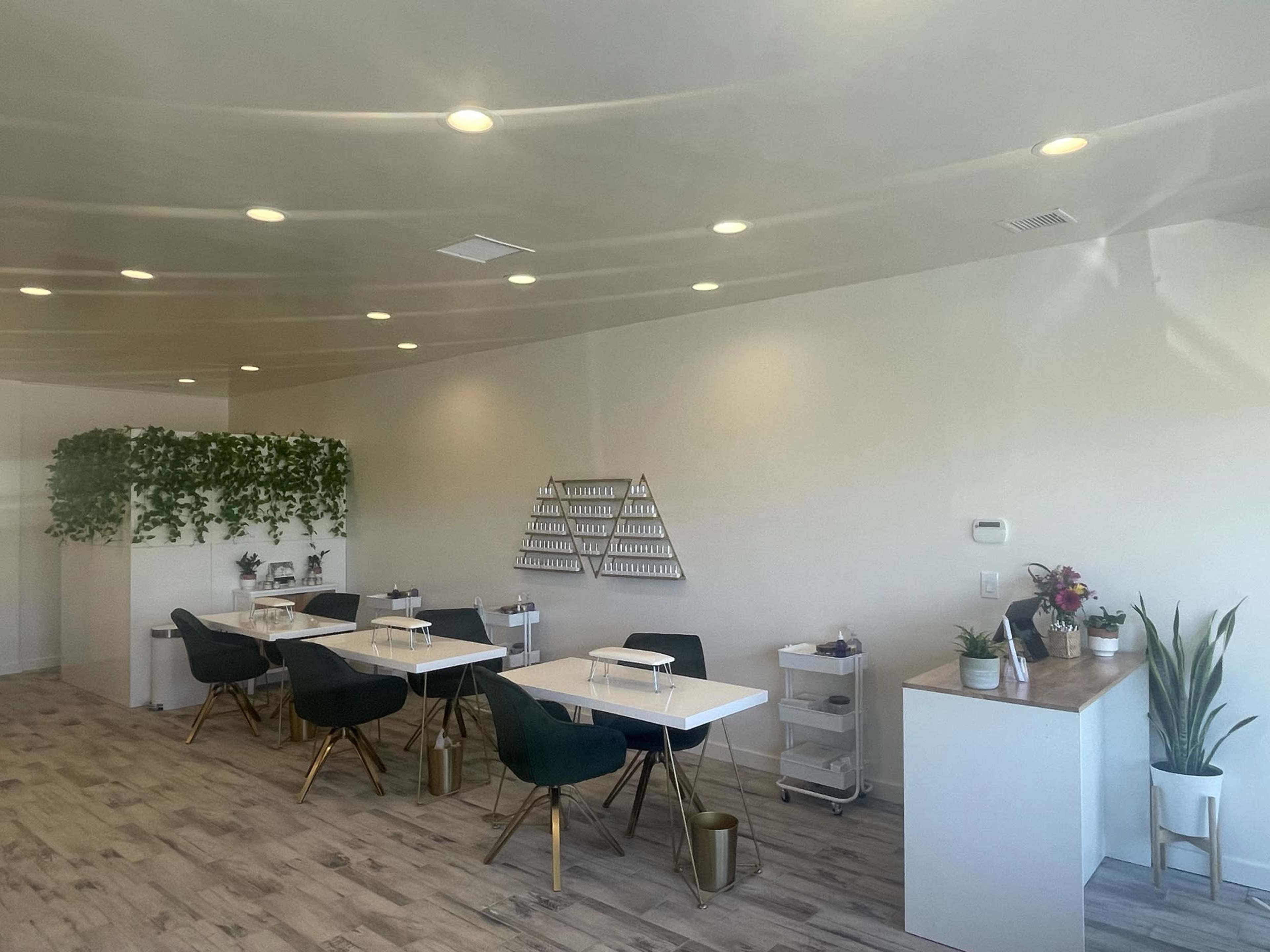 A modern, minimalistic interior of a nail salon with white walls, several treatment stations with chairs, and a partition adorned with greenery.