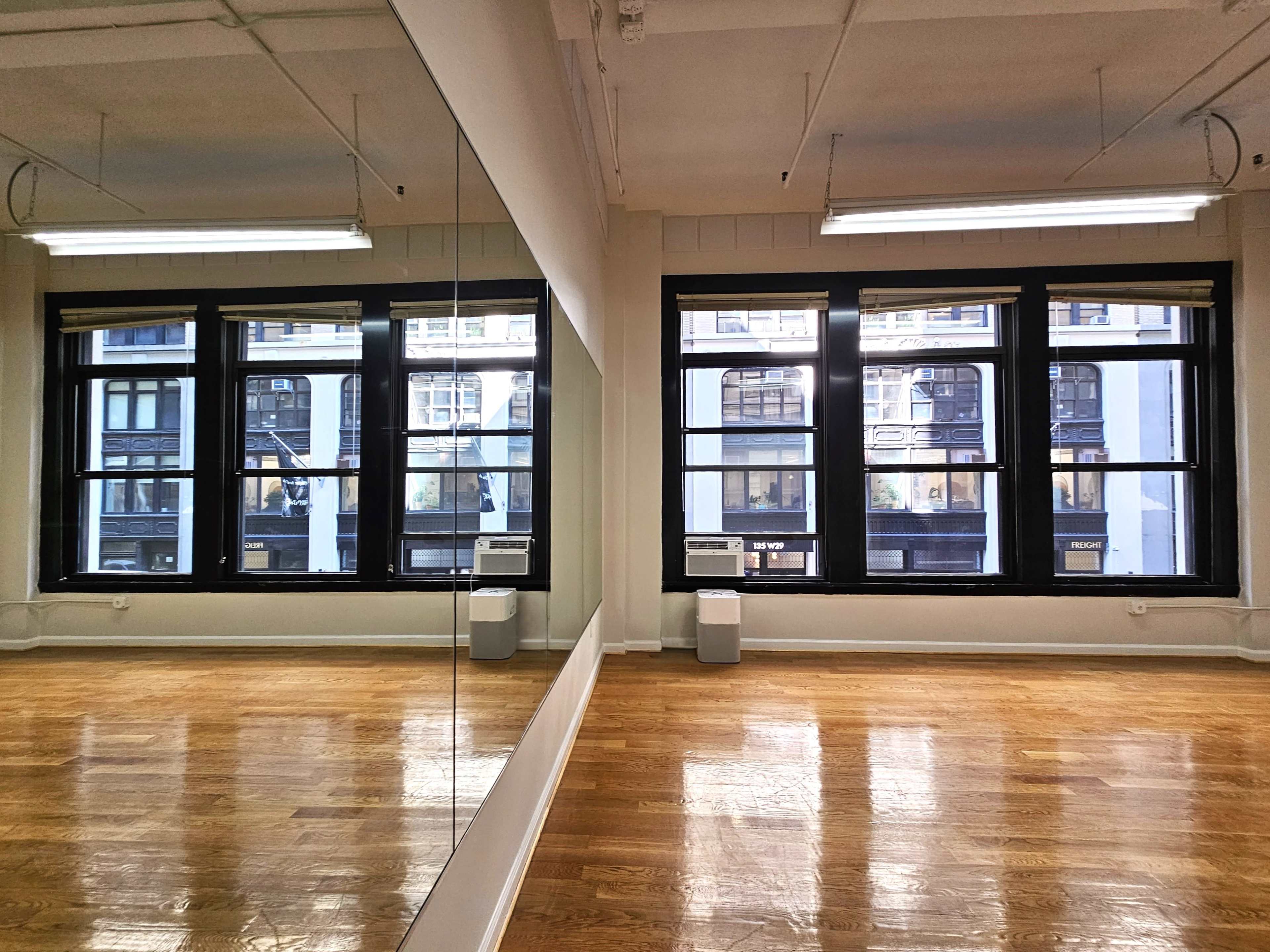 The image shows a spacious room with polished wooden floors, large windows on one side that overlook a street, and a full-length mirror mounted on the opposite wall.