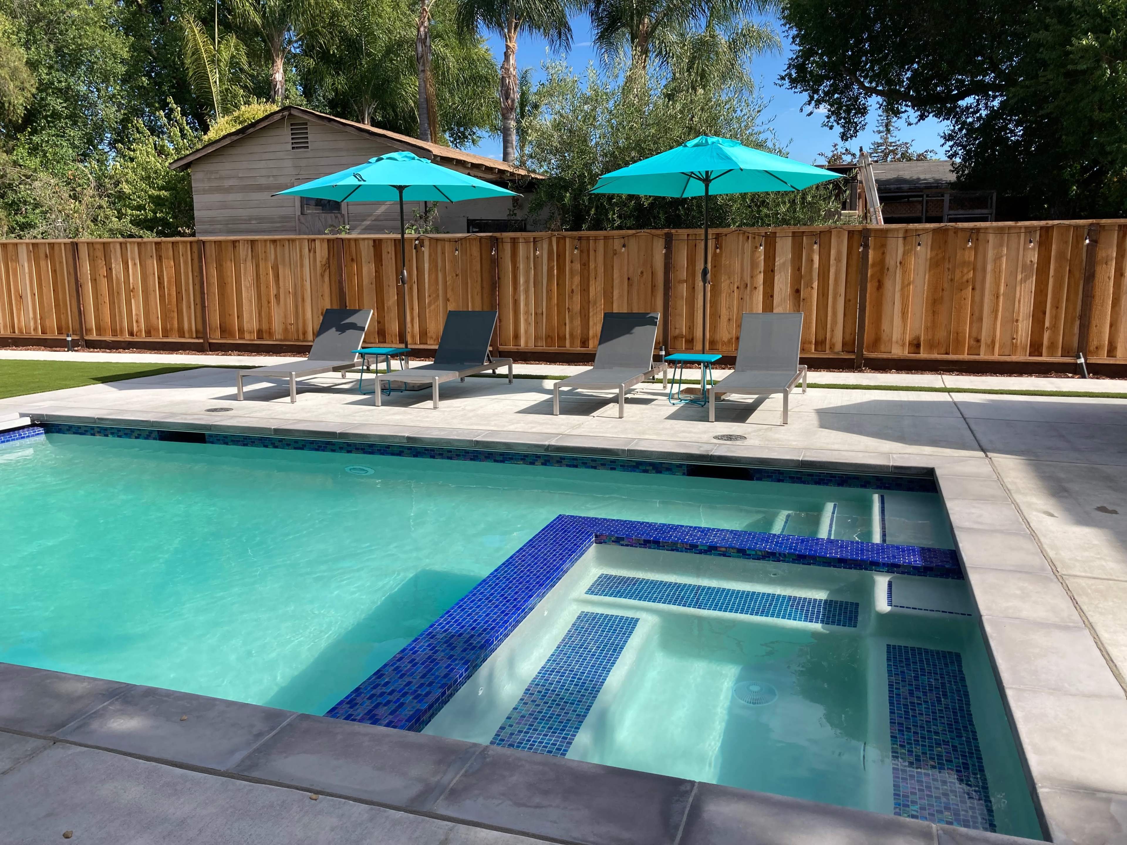 A swimming pool with a spa area, surrounded by four lounge chairs and two teal umbrellas, with a wooden fence and palm trees in the background.