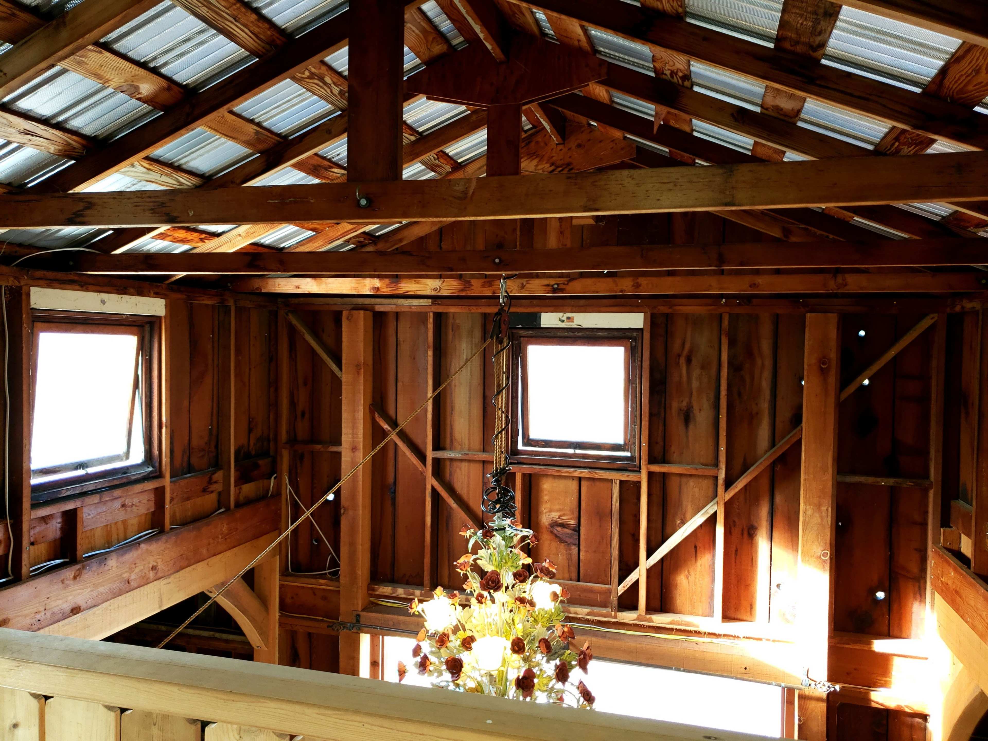 The interior of a wooden structure with exposed beams, two windows, and a chandelier hanging from the ceiling.