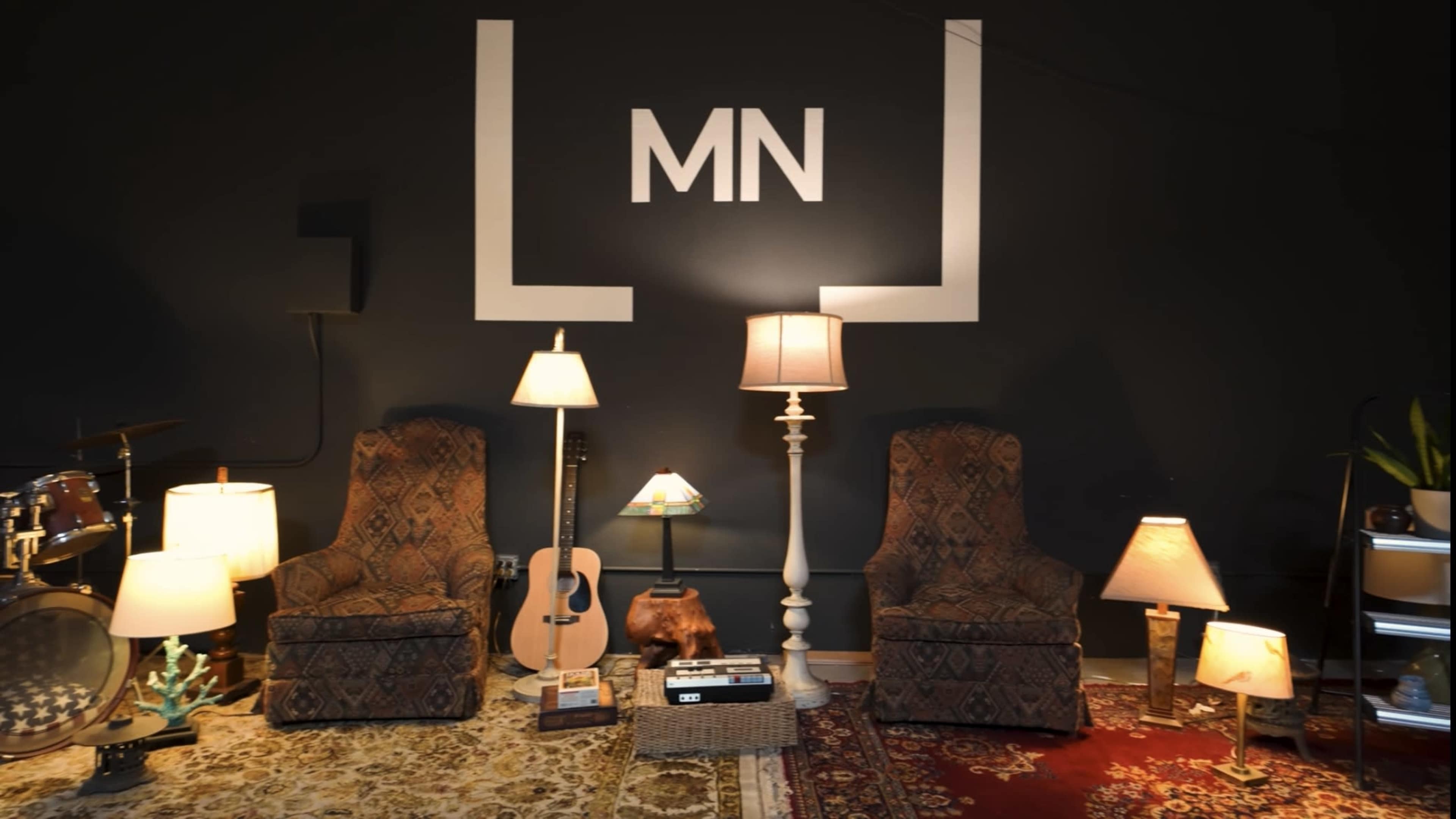 The scene features two patterned armchairs flanking a lamp on a patterned rug, with an acoustic guitar and decorative items against a dark wall marked with "MN."