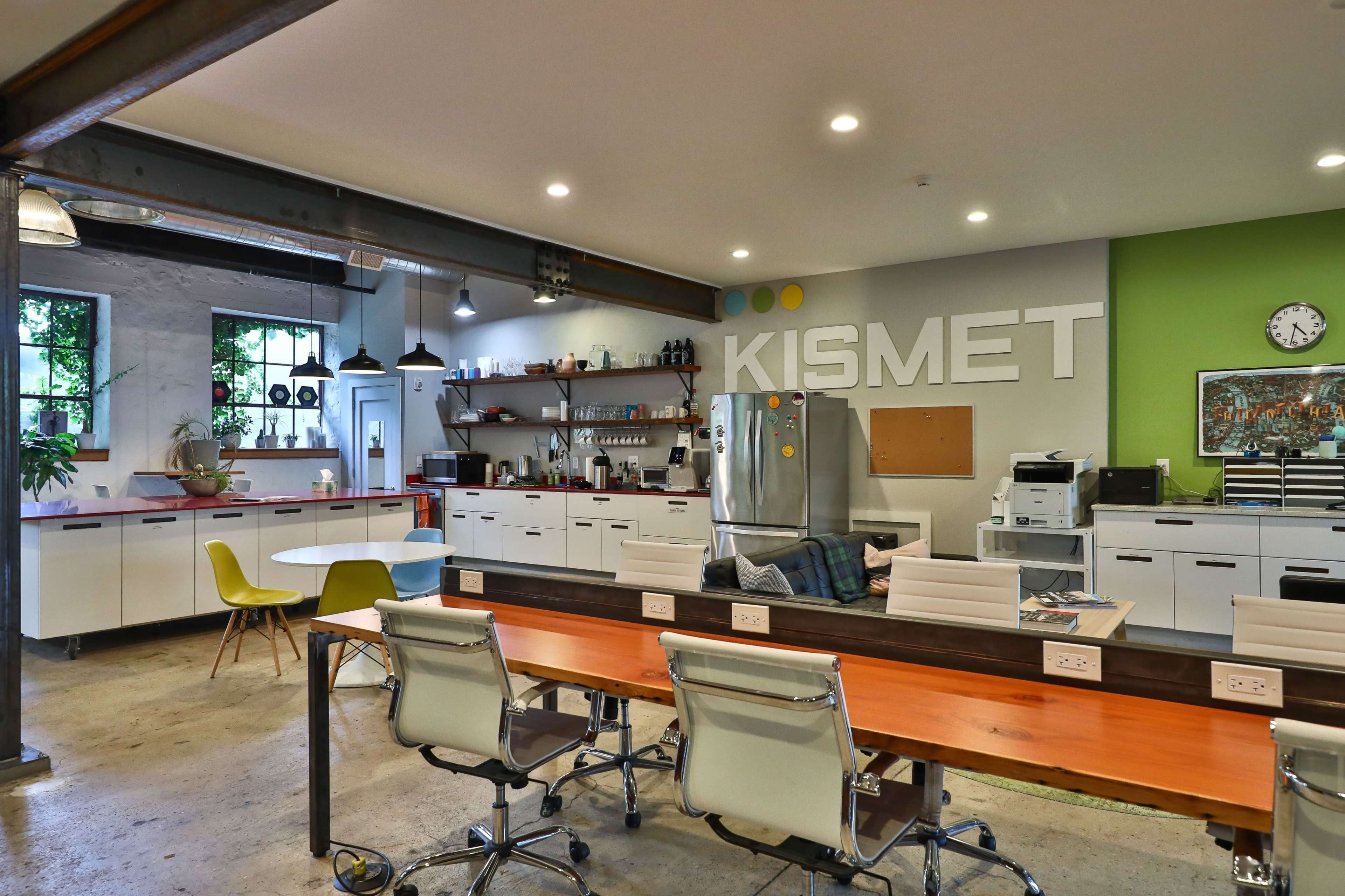 A modern office kitchen area featuring a communal table, chairs, a refrigerator, and shelves with various kitchen supplies, all adorned with the word "KISMET" on the wall.