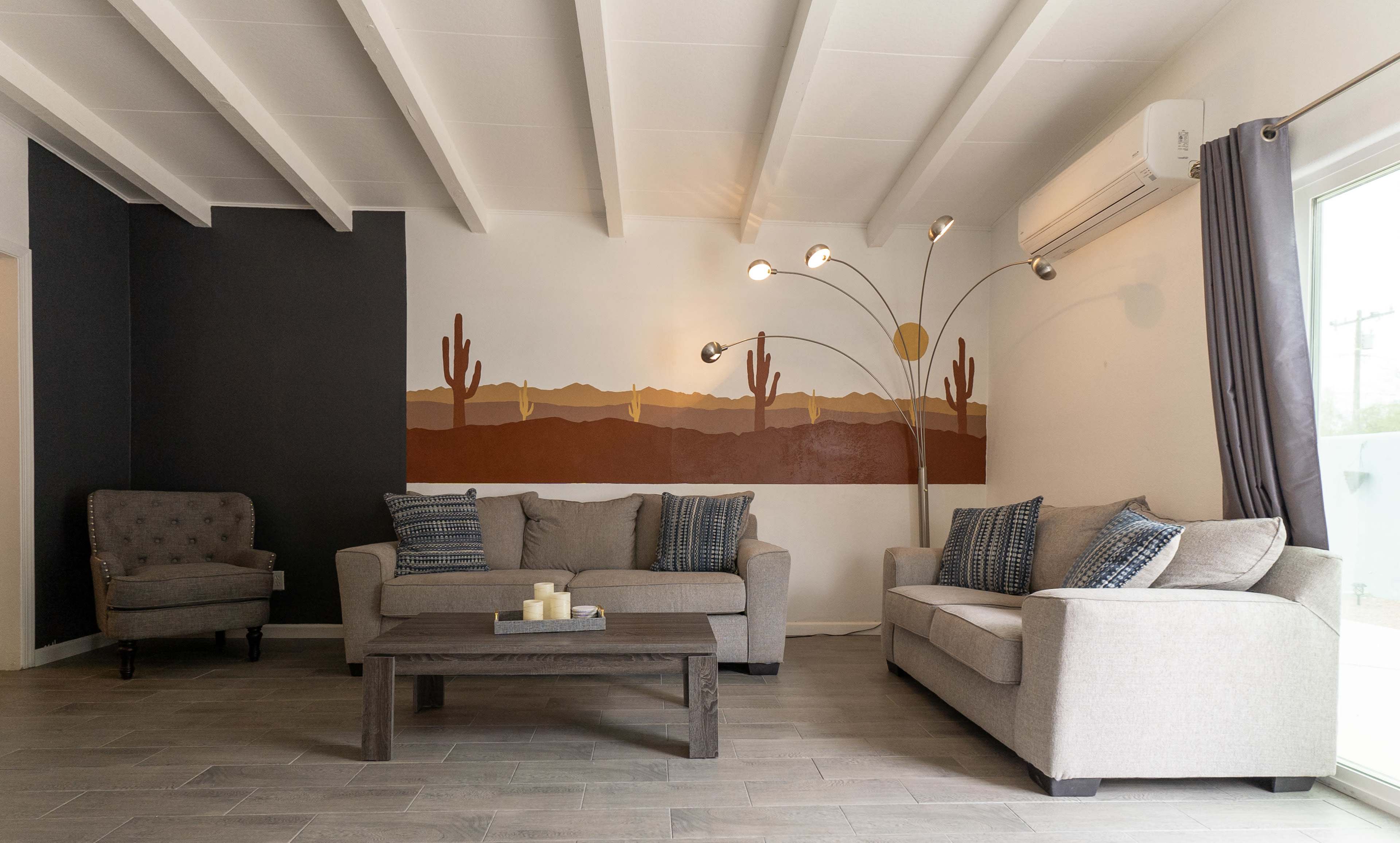 A modern living room features two light gray sofas, a wooden coffee table, and a decorative mural of cacti and mountains on the wall.