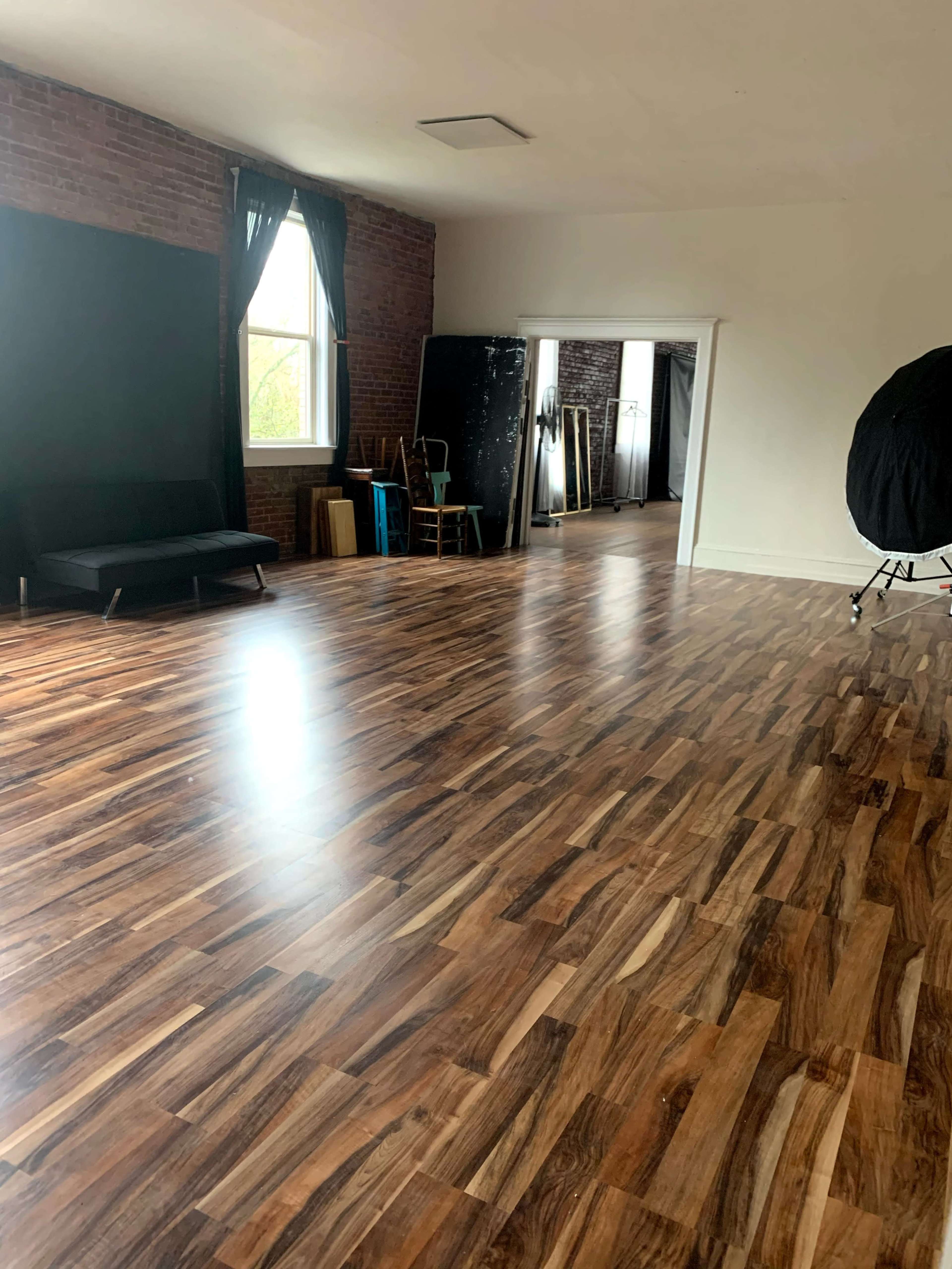 A spacious room with wooden flooring, large windows, and a simple seating area, alongside various studio equipment in the background.