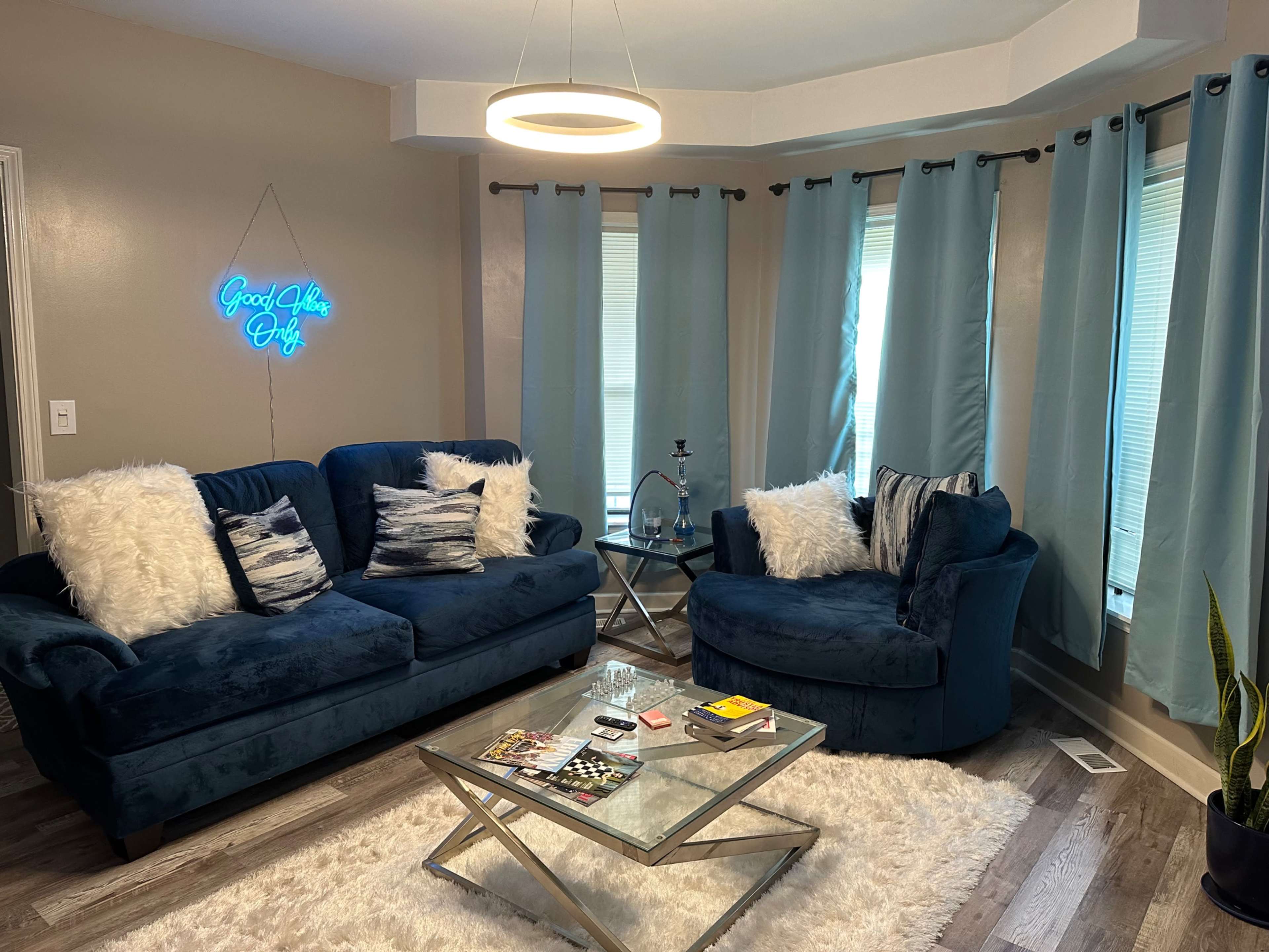 The living room features two blue sofas with decorative pillows, a glass coffee table, and large windows covered with light blue curtains.