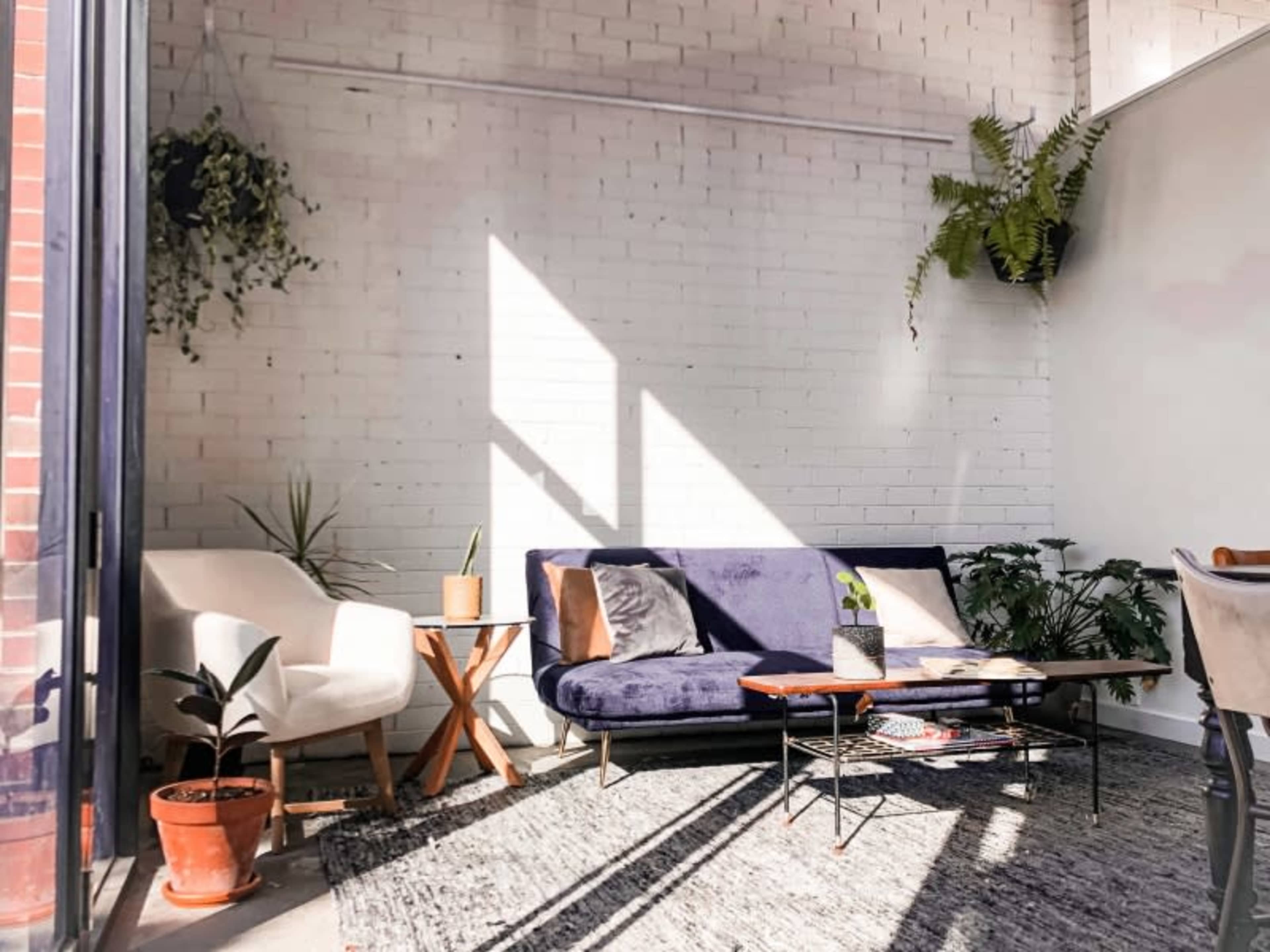 A cozy living area features a purple sofa, a white armchair, a wooden coffee table, and various plants against a bright, white brick wall.