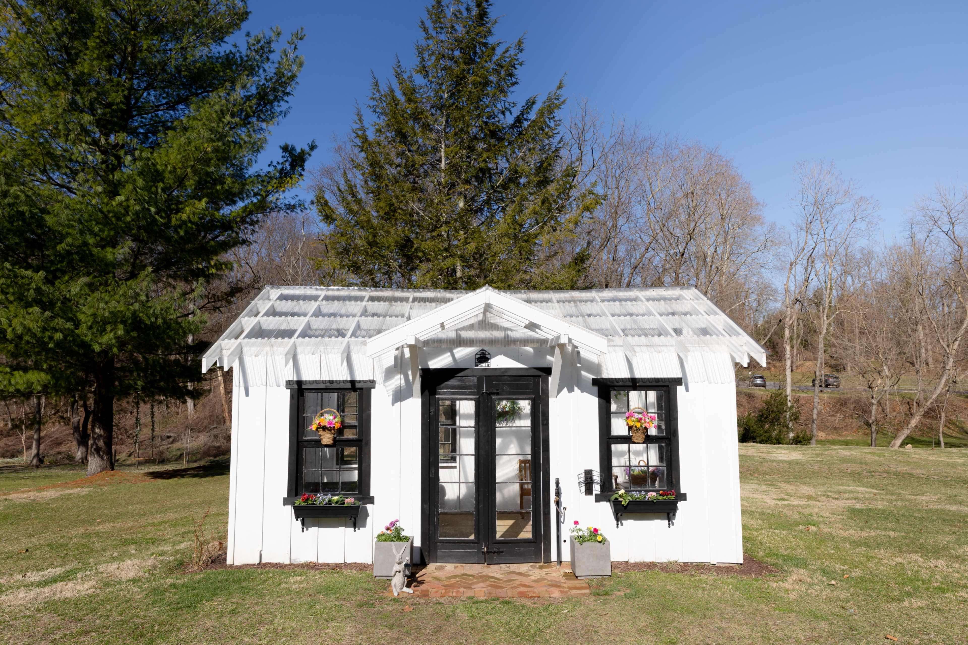 The image shows a small white building with a transparent roof, black-framed windows, and flower boxes, surrounded by a grassy area and trees.