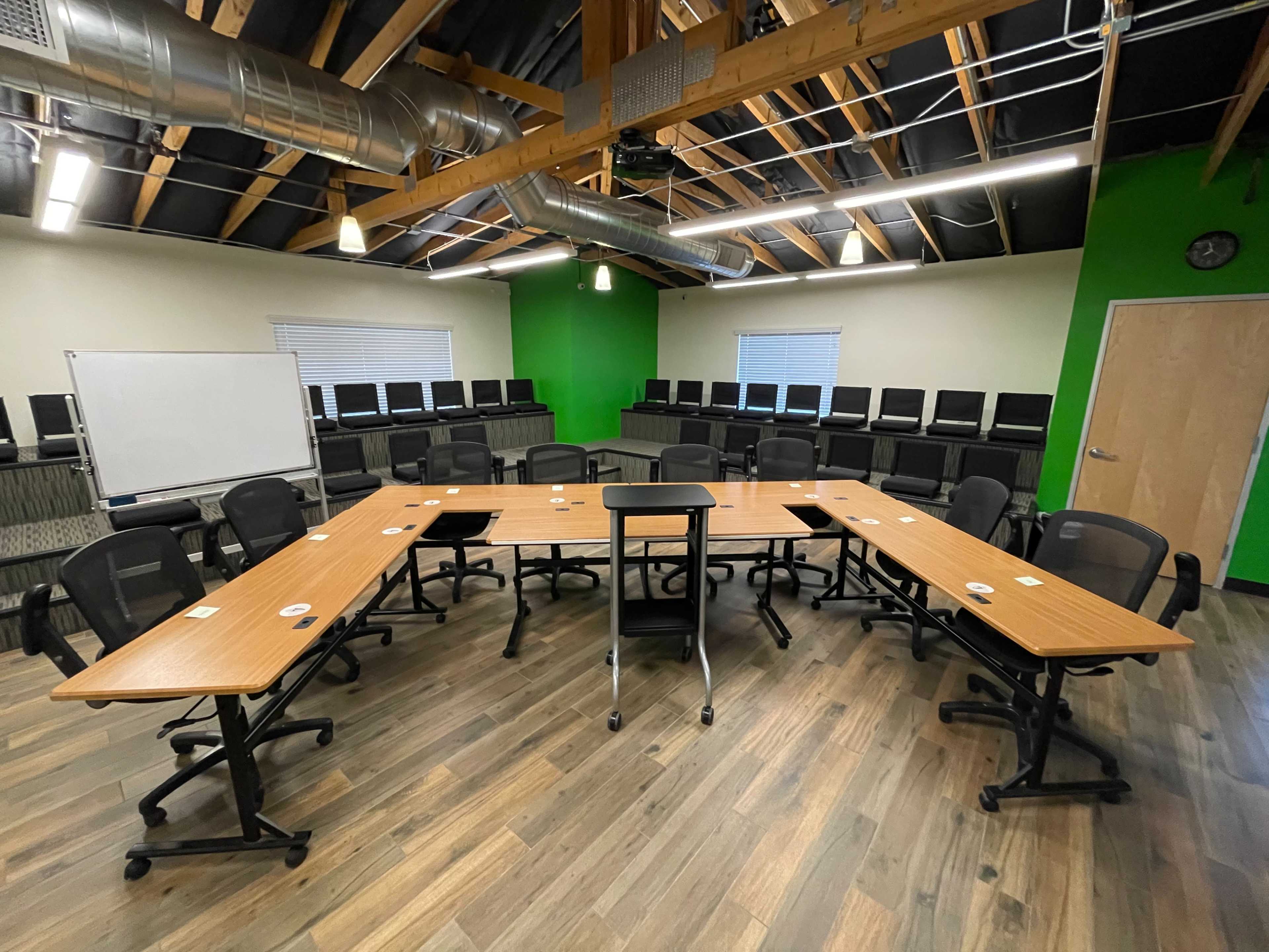 A large conference room features a U-shaped arrangement of wooden tables surrounded by black chairs, with a whiteboard and multiple rows of chairs against the back wall.