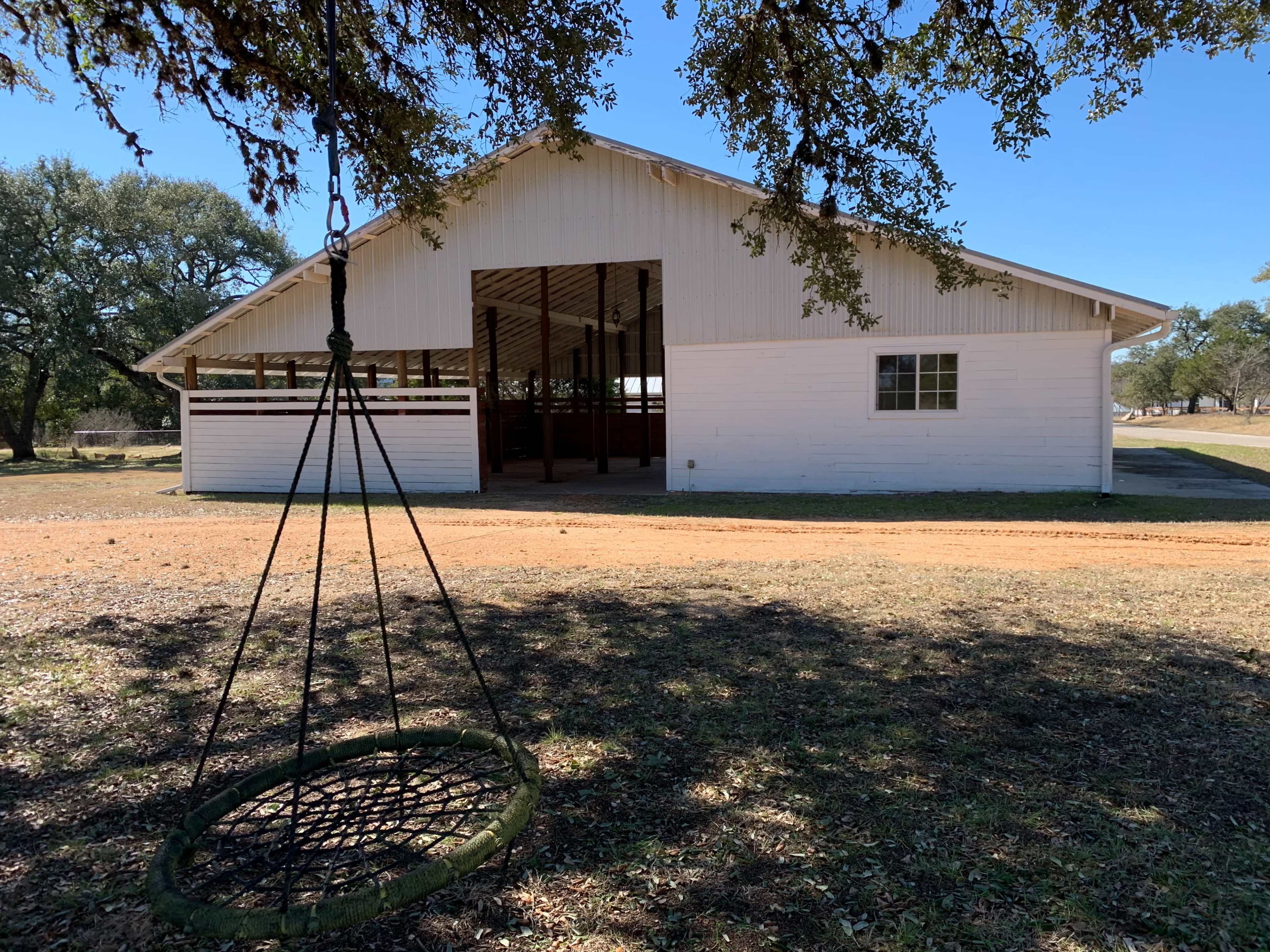 A white barn with large open doors stands beside a dirt path, and a hanging swing made of ropes and a circular seat is in the foreground.