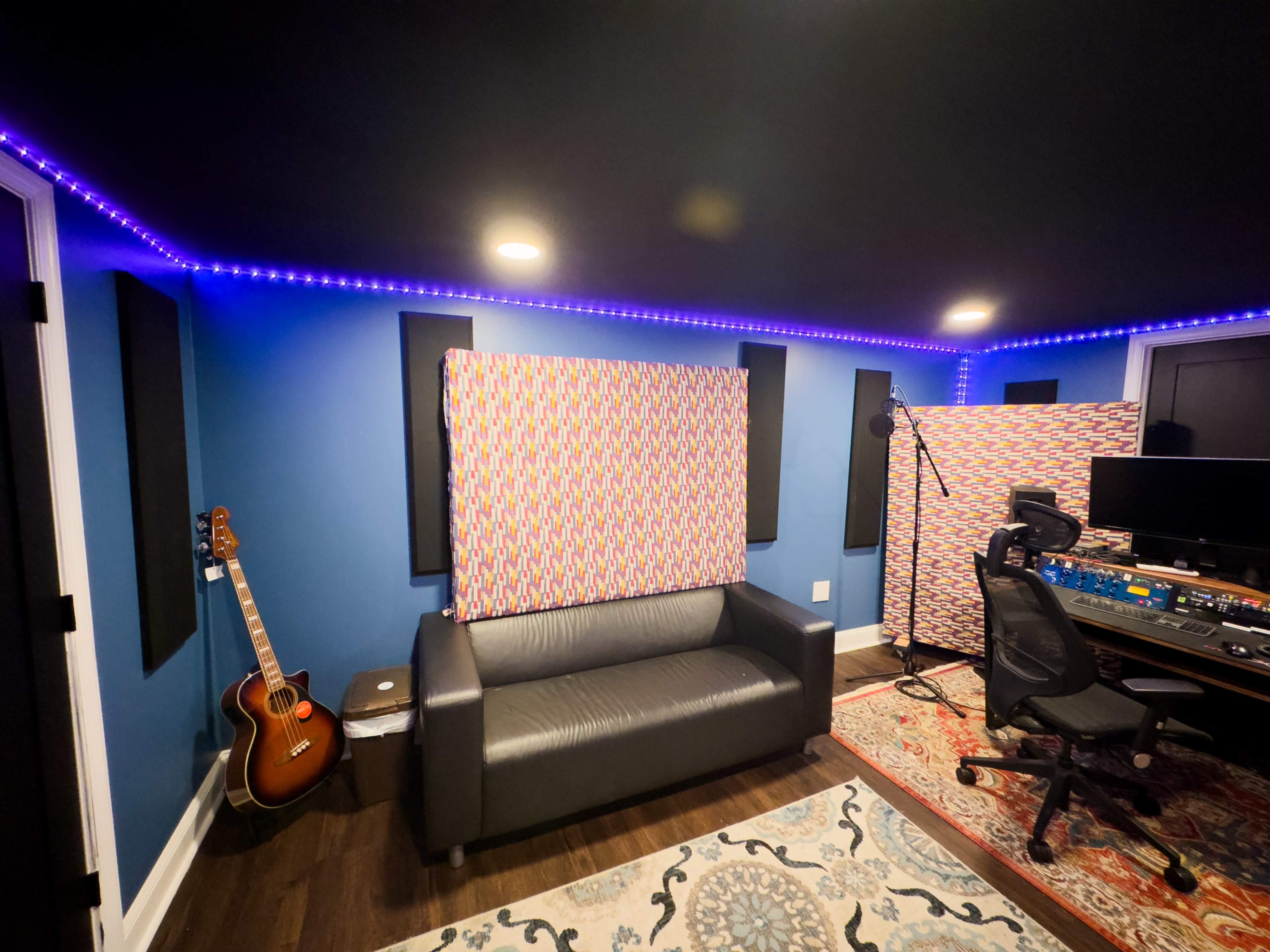 A recording studio featuring a black couch, acoustic panels on the walls, a guitar in the corner, and a mixing console with a swivel chair.