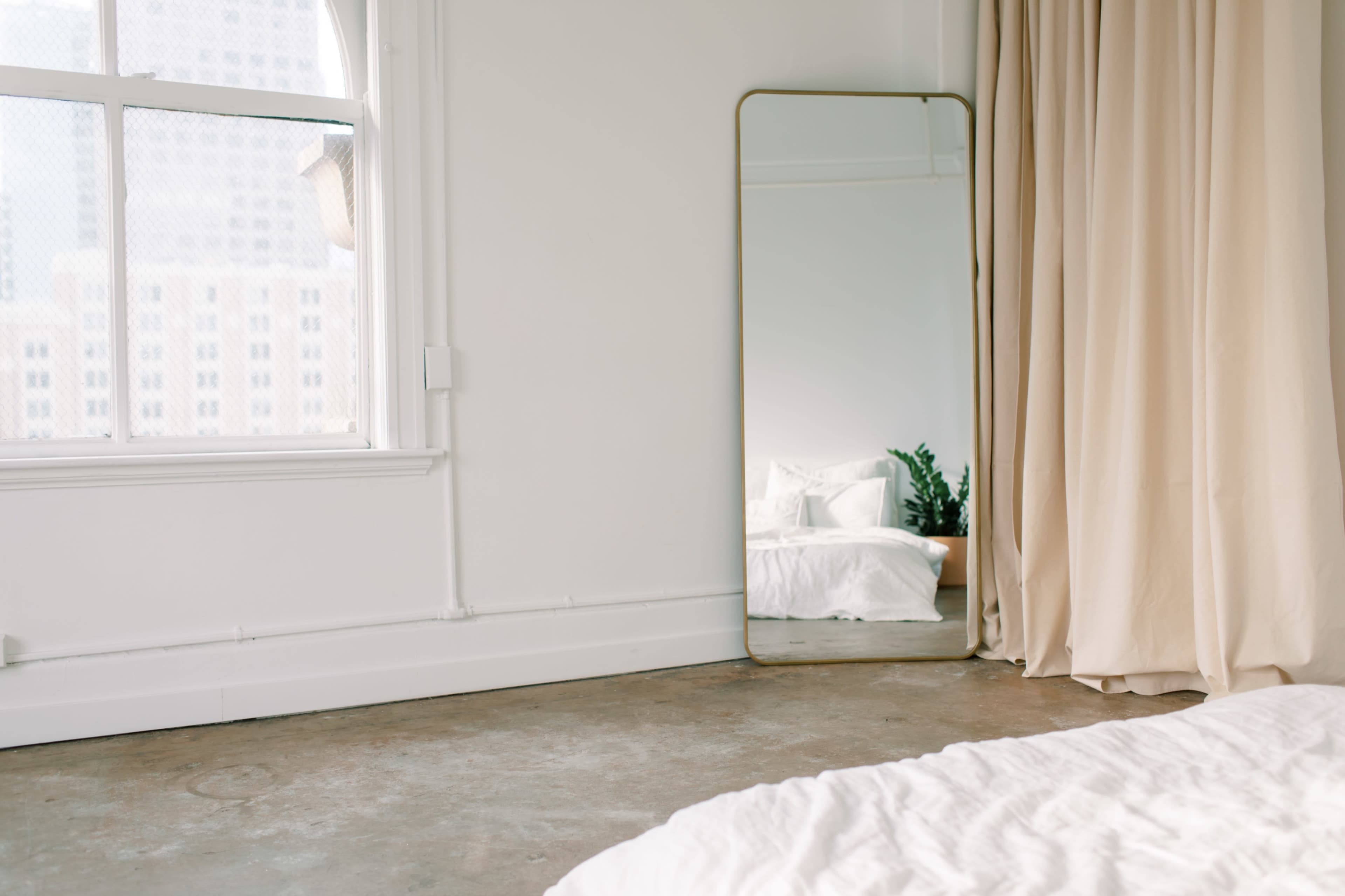 A minimalist bedroom with a large mirror next to a window, and a bed with white linens in the foreground.