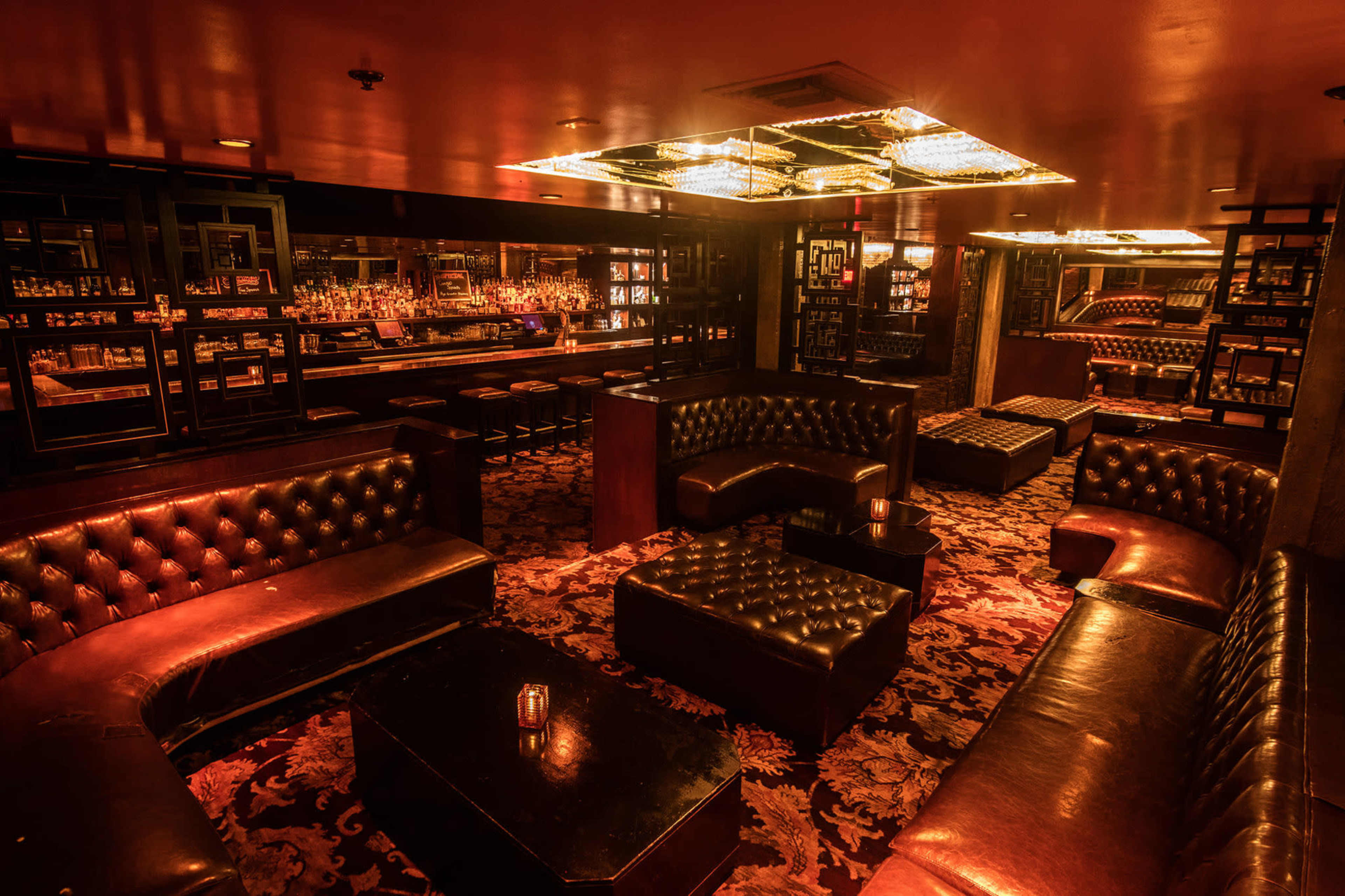 The image shows a dimly lit lounge with dark leather seating, a bar area, and an ornate ceiling fixture.